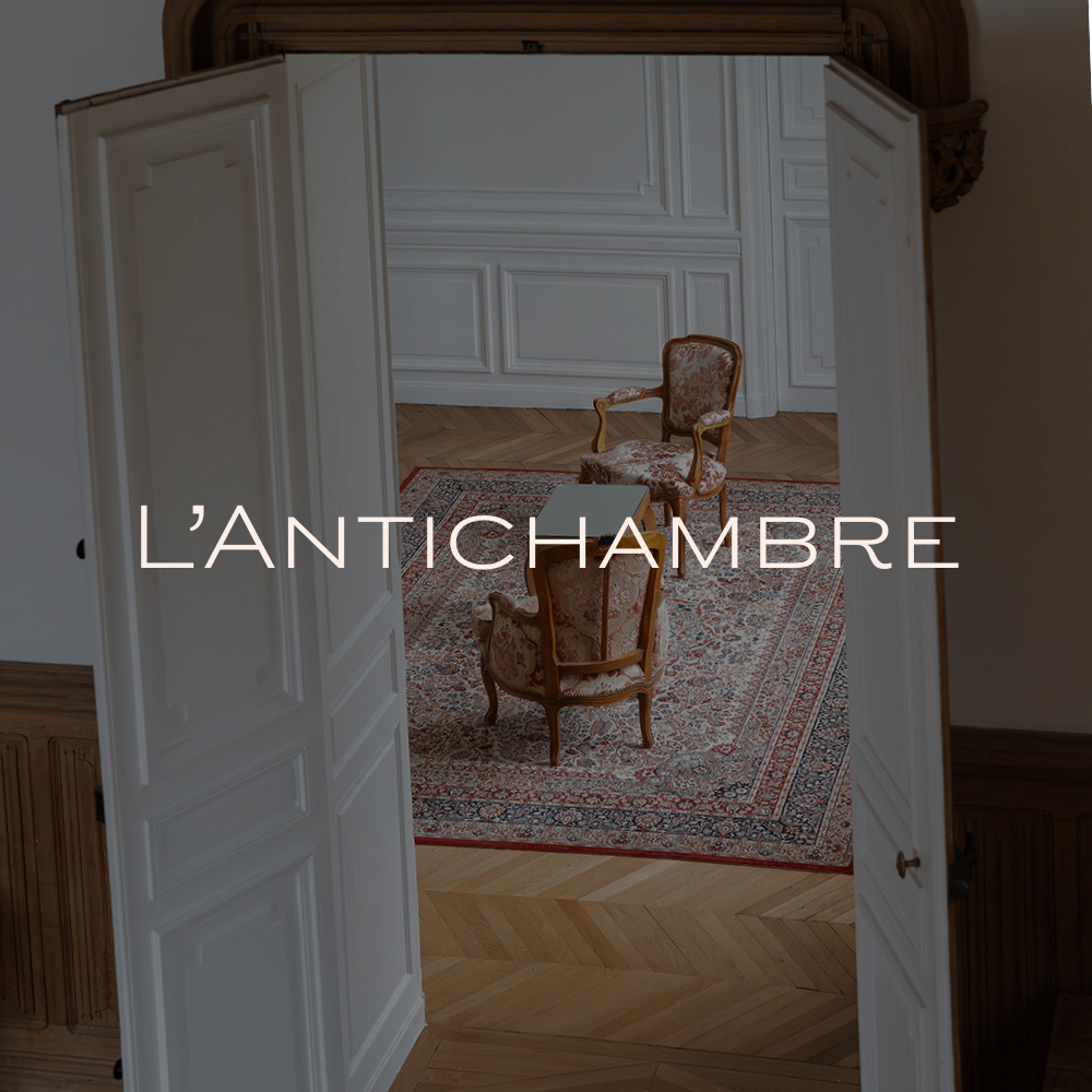 Une pièce appelée 'L'Antichambre' d'une superficie de 25 mètres carrés, décorée avec une table ancienne, un gâteau décoratif, un bouquet de fleurs, et un seau à champagne en métal.