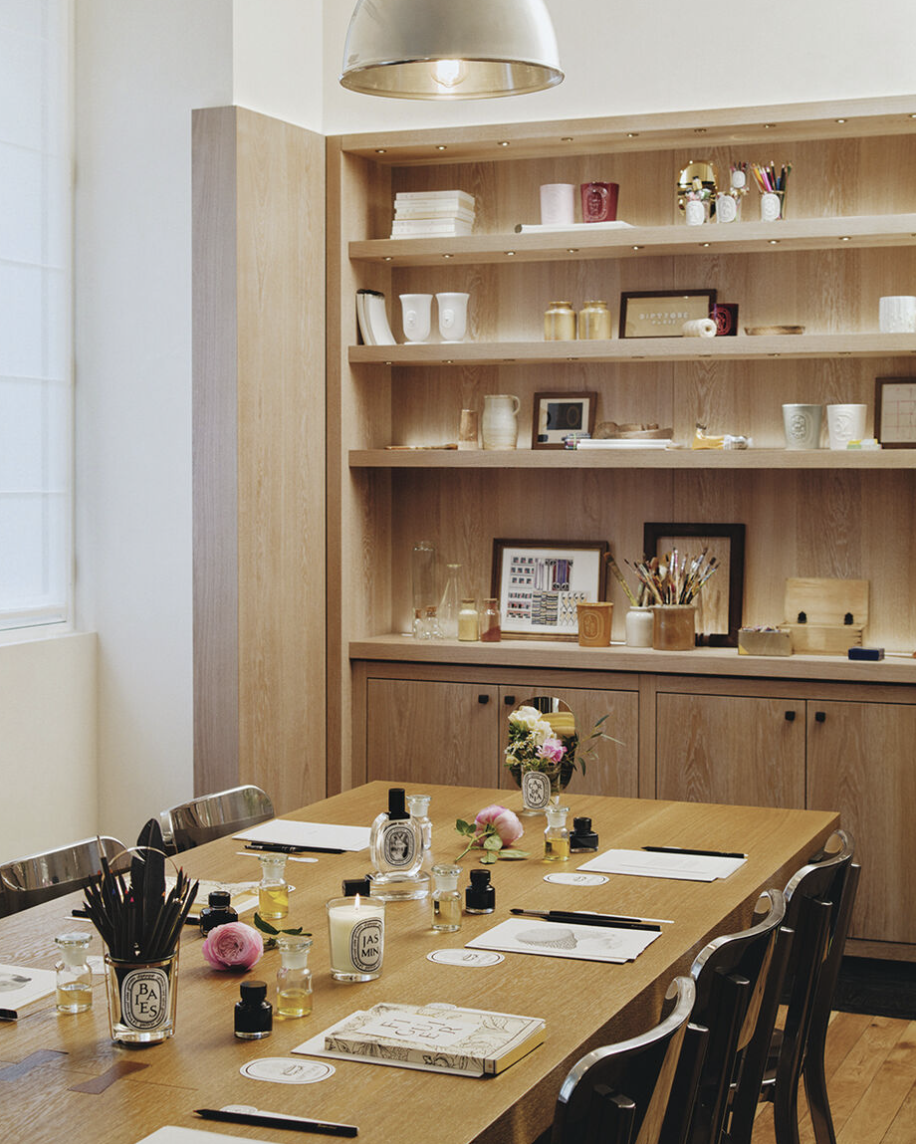 Une salle de réunion avec une grande table en bois, entourée de chaises, avec des fiches, des stylos, des bouteilles de parfum ou d'huiles essentielles, et des fleurs. En arrière-plan, une grande étagère en bois présente des objets décoratifs, des ca