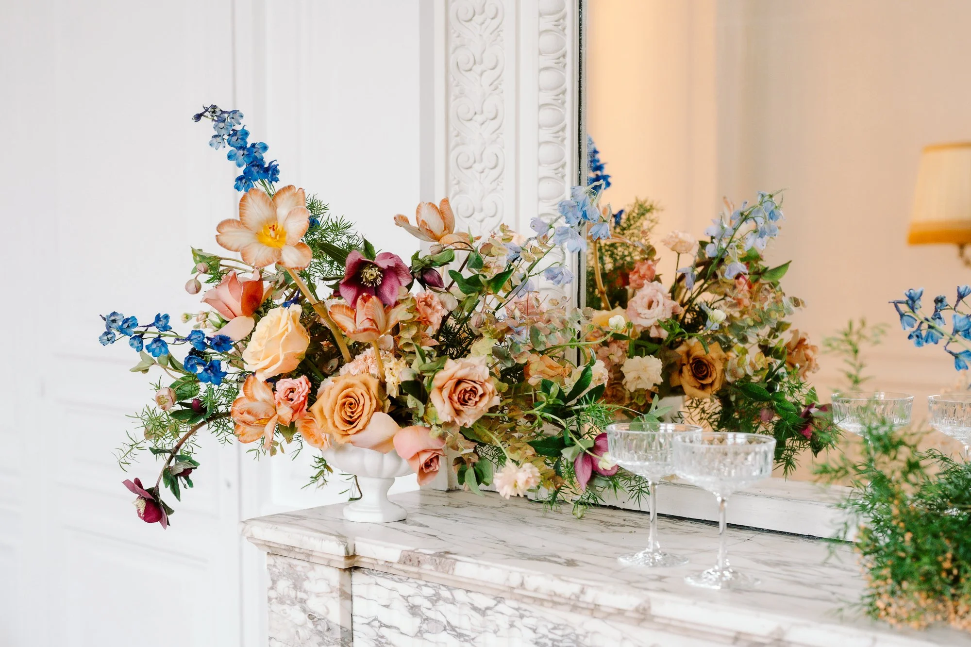 Grand bouquet de fleurs colorées dans un vase blanc sur une console en marbre, reflété dans un miroir au mur, avec quelques verres en cristal à côté.