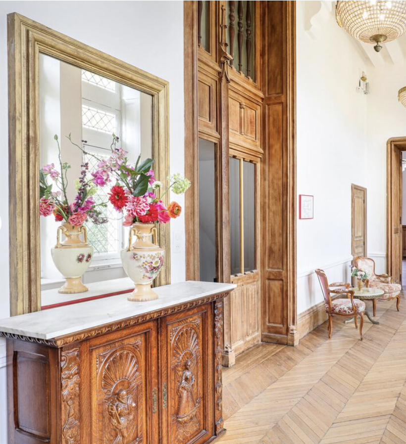 Intérieur d'une pièce avec un meuble en bois sculpté, un miroir, des vases avec des fleurs, deux chaises avec des coussins, un plafond en bois, un parquet en bois et des lampes en papier.