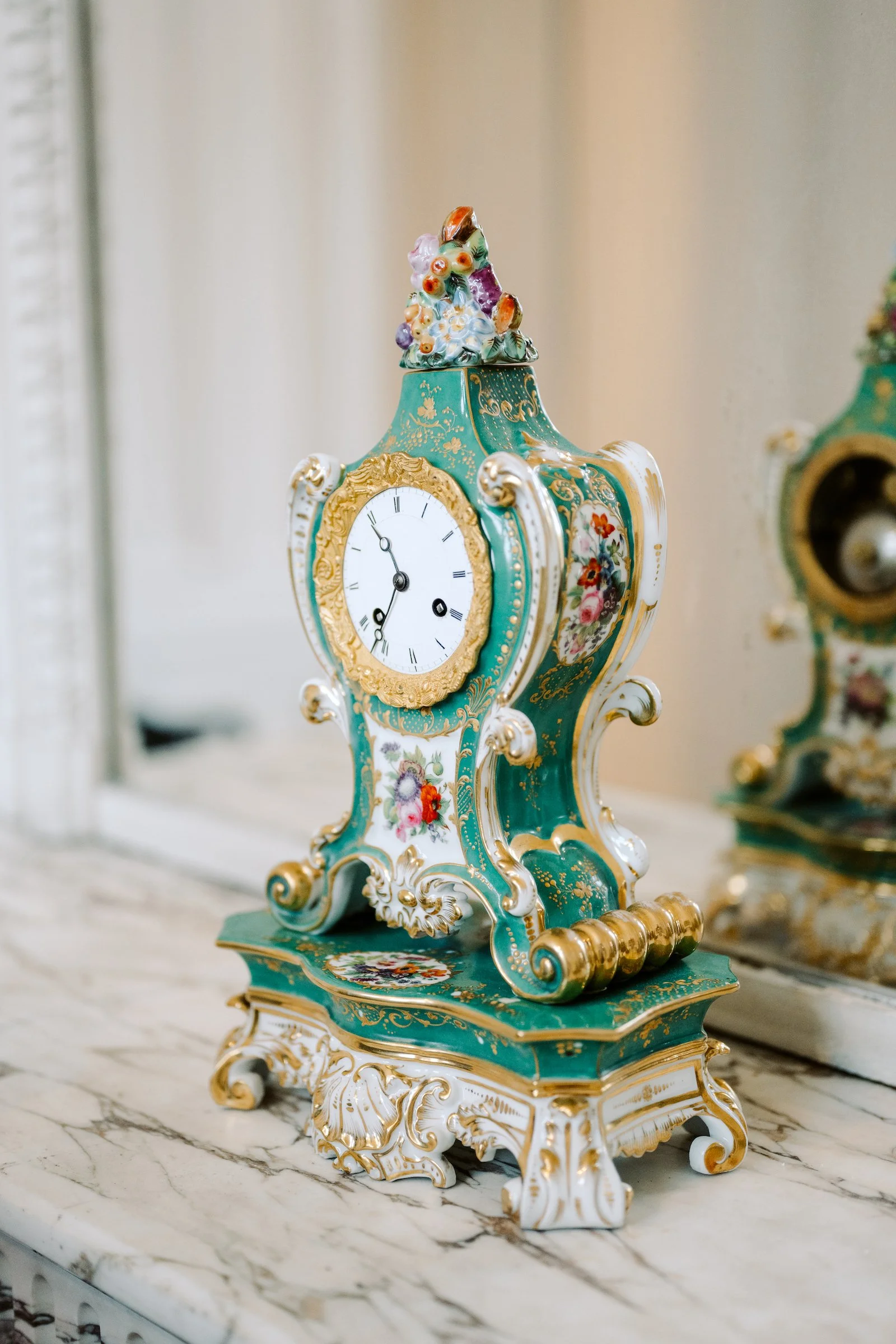 Une horloge ornementale en porcelaine de style rococo avec des détails dorés, décorée de motifs floraux, posée sur une surface en marbre.