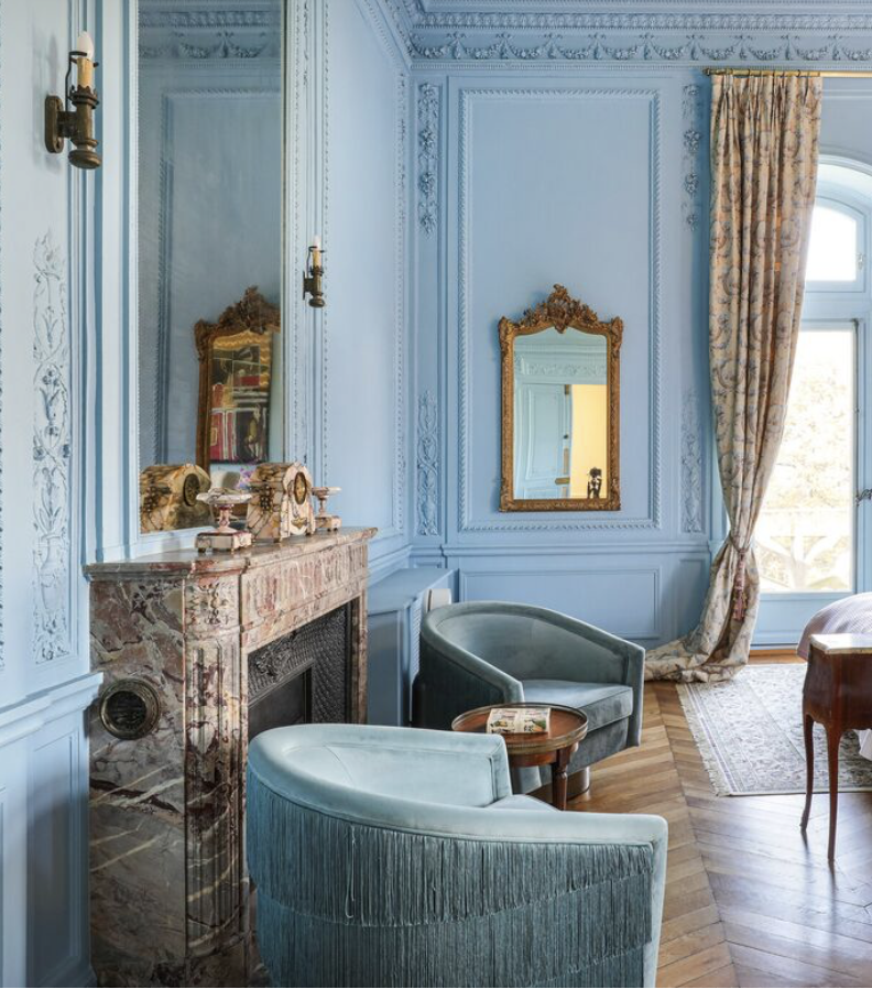Intérieur d'une pièce élégante avec murs bleu clair ornés de moulures, un miroir doré, un feu de cheminée en marbre, deux fauteuils en velours vert menthe, une petite table en bois avec un livre, une grande fenêtre avec des rideaux à motifs.