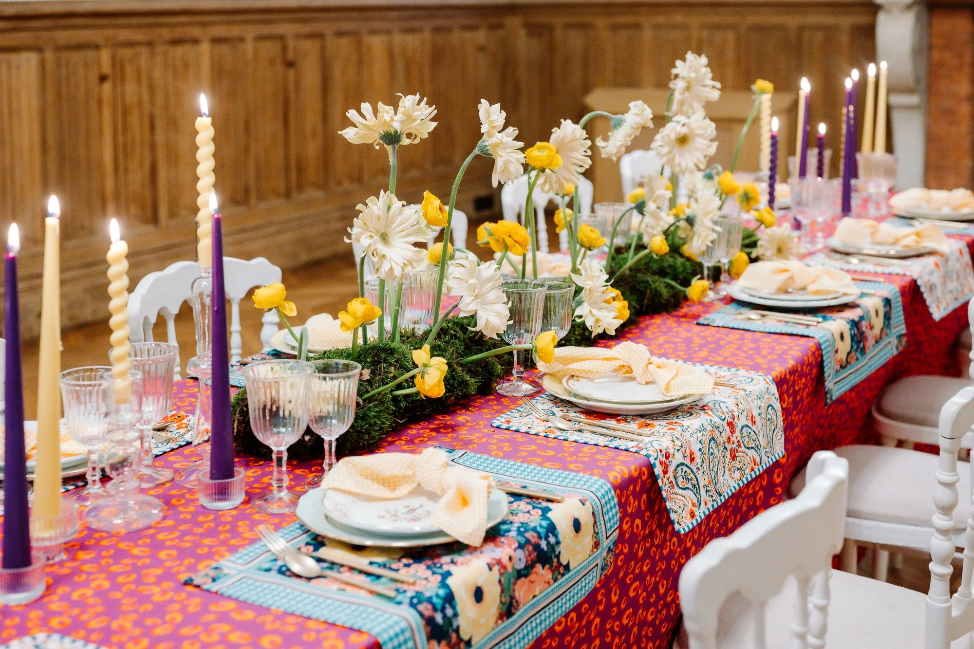 Table magnifiquement dressée avec des fleurs blanches et jaunes en centre, des bougies torsadées violettes et jaunes, des verres élégants, des assiettes avec des serviettes pliées, avec un décor coloré de nappe et de sets de table multicolores, dans