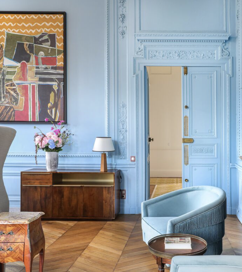 Salle élégante avec murs bleus, cheminée ornée, porte en bois coloré, tableau moderne, vase de fleurs, lampe de table, fauteuil en tissu bleu avec un magazine, parquet en bois clair.