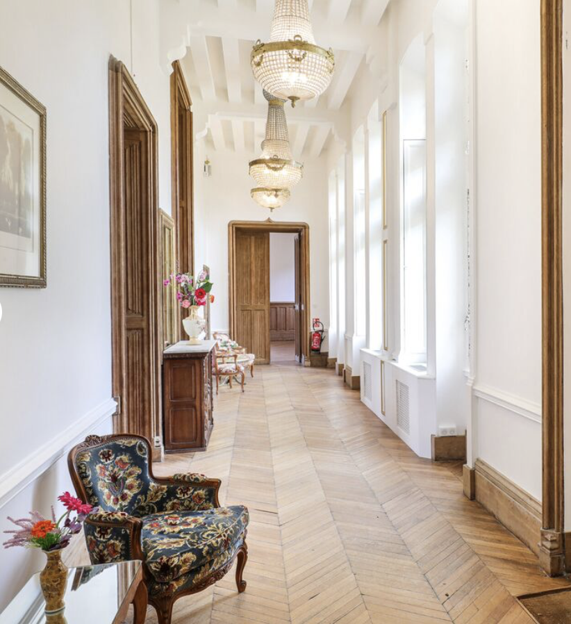 Couloir élégant avec parquet en bois, luminaires en verre suspendus, chaises et un fauteuil ancien, fleurs dans un vase, porte en bois et fenêtres lumineuses.