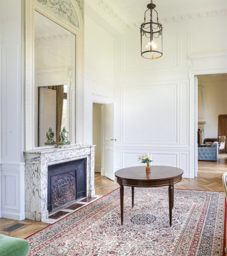 Un salon élégant avec un tapis orientale, une cheminée en marbre, un miroir grand et une table ronde avec un vase de fleurs, et un lustre suspendu.