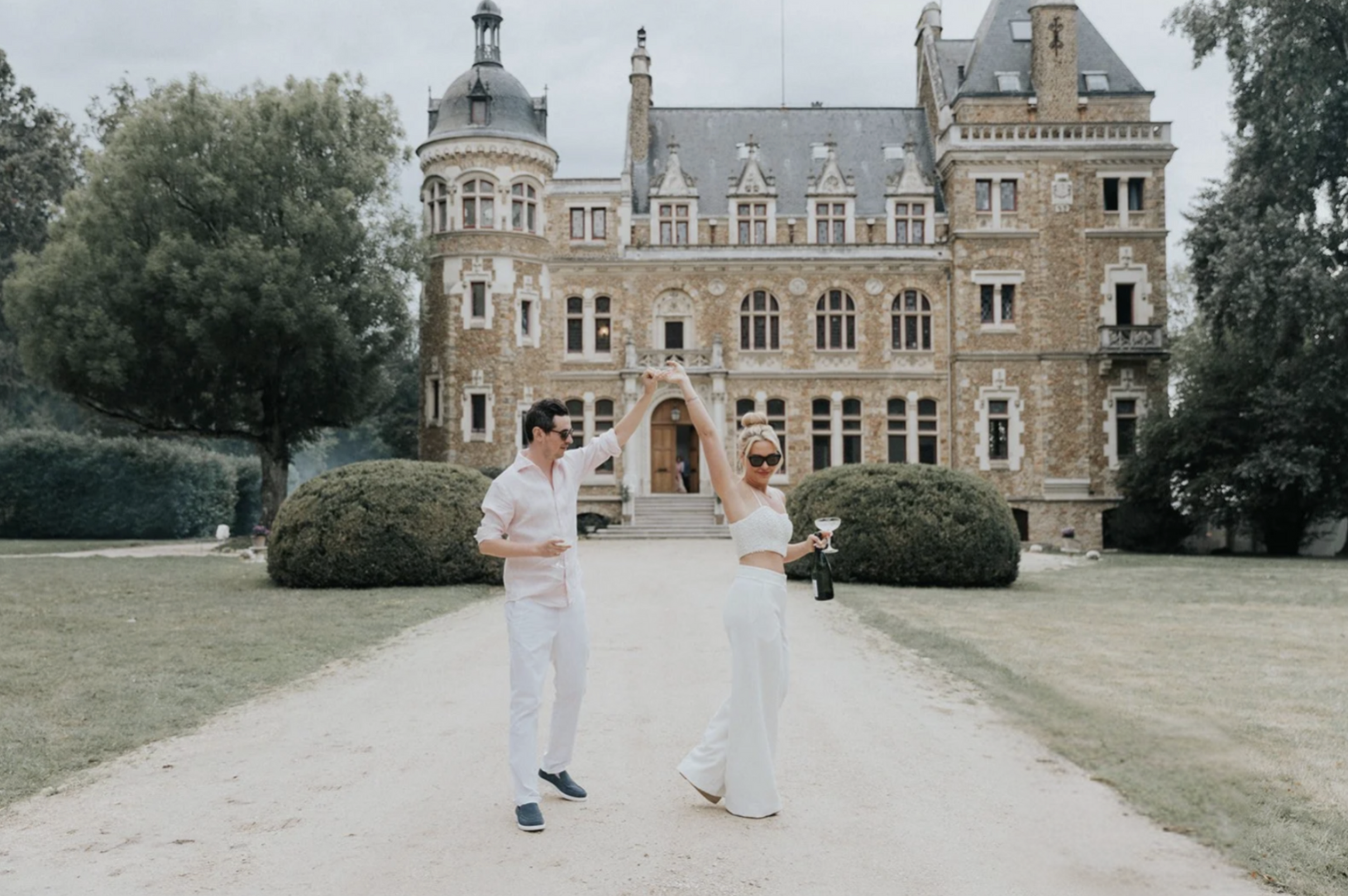 Un couple danse devant un château historique entouré d'arbres et de buissons, la femme tient un verre de cocktail et une bouteille de champagne, ils portent des vêtements d'été élégants.