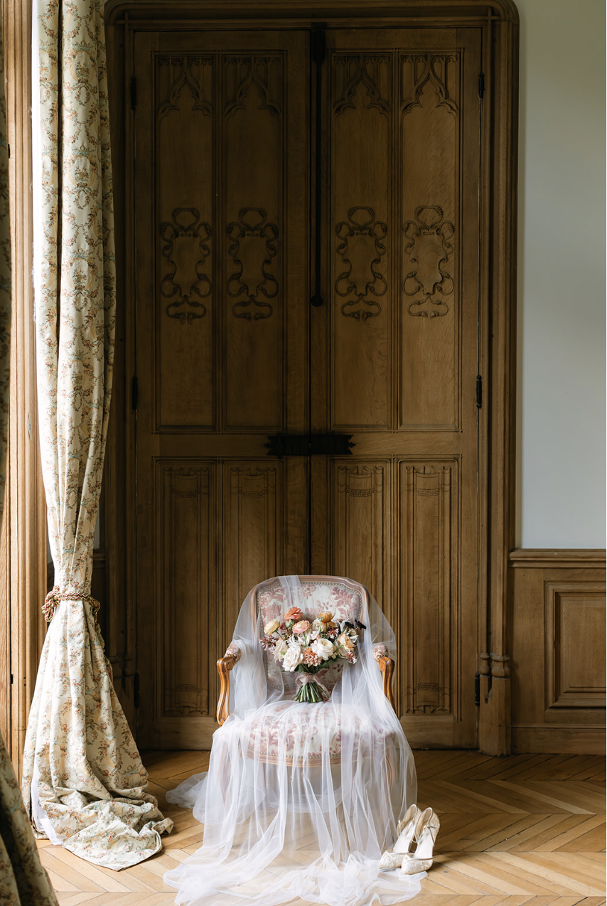 Une chaise ancienne en bois avec un coussin, recouverte d'un voile blanc, portant un bouquet de fleurs roses, blanches et orange, avec une paire de chaussures de mariage en dessous. La pièce a un sol en bois et des rideaux fleuris à gauche, avec un g