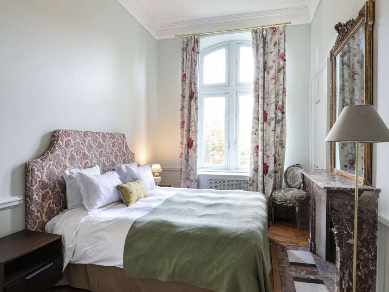 Chambre élégante avec lit double, tête de lit ornée, rideaux fleuris, grande fenêtre laissant entrer la lumière naturelle, miroir ancien, lampe de chevet, table en marbre, chaise ancienne, tapis au sol.