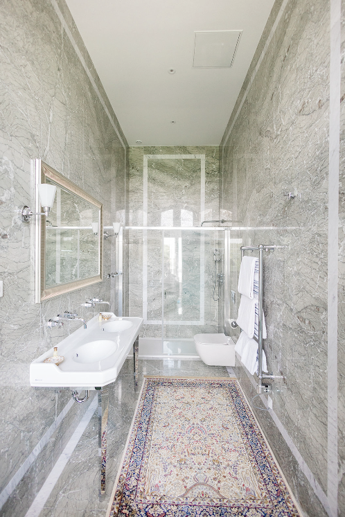 Salle de bain moderne avec murs en marbre, lavabo blanc, grand miroir, douche avec parois en verre, bidet, porte-serviettes chauffant avec serviettes, tapis persan au sol.
