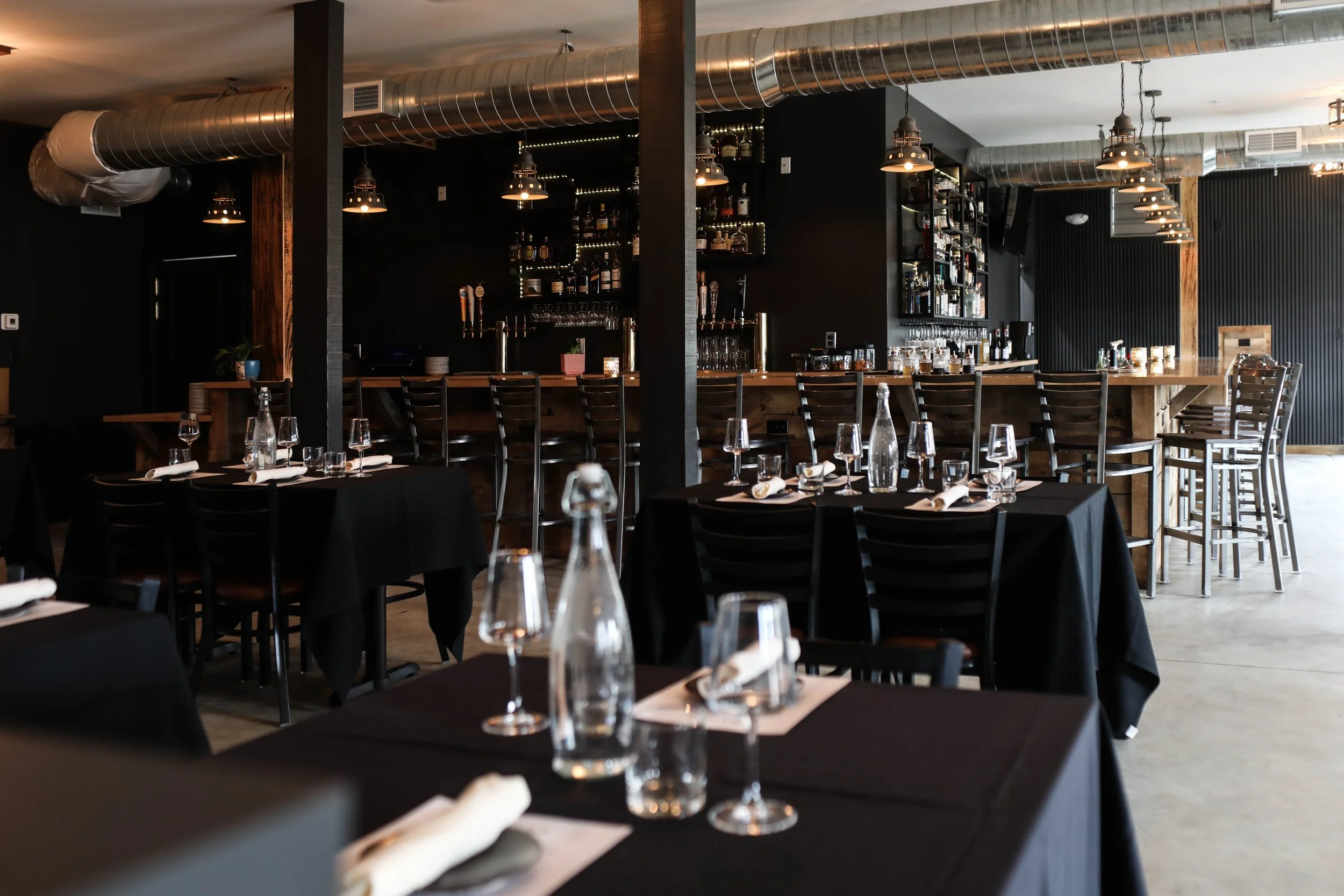 Upscale restaurant dining area with black tablecloths, glassware, and cutlery, overlooking a bar with bottles and wine glasses, industrial ventilation pipes on ceiling.