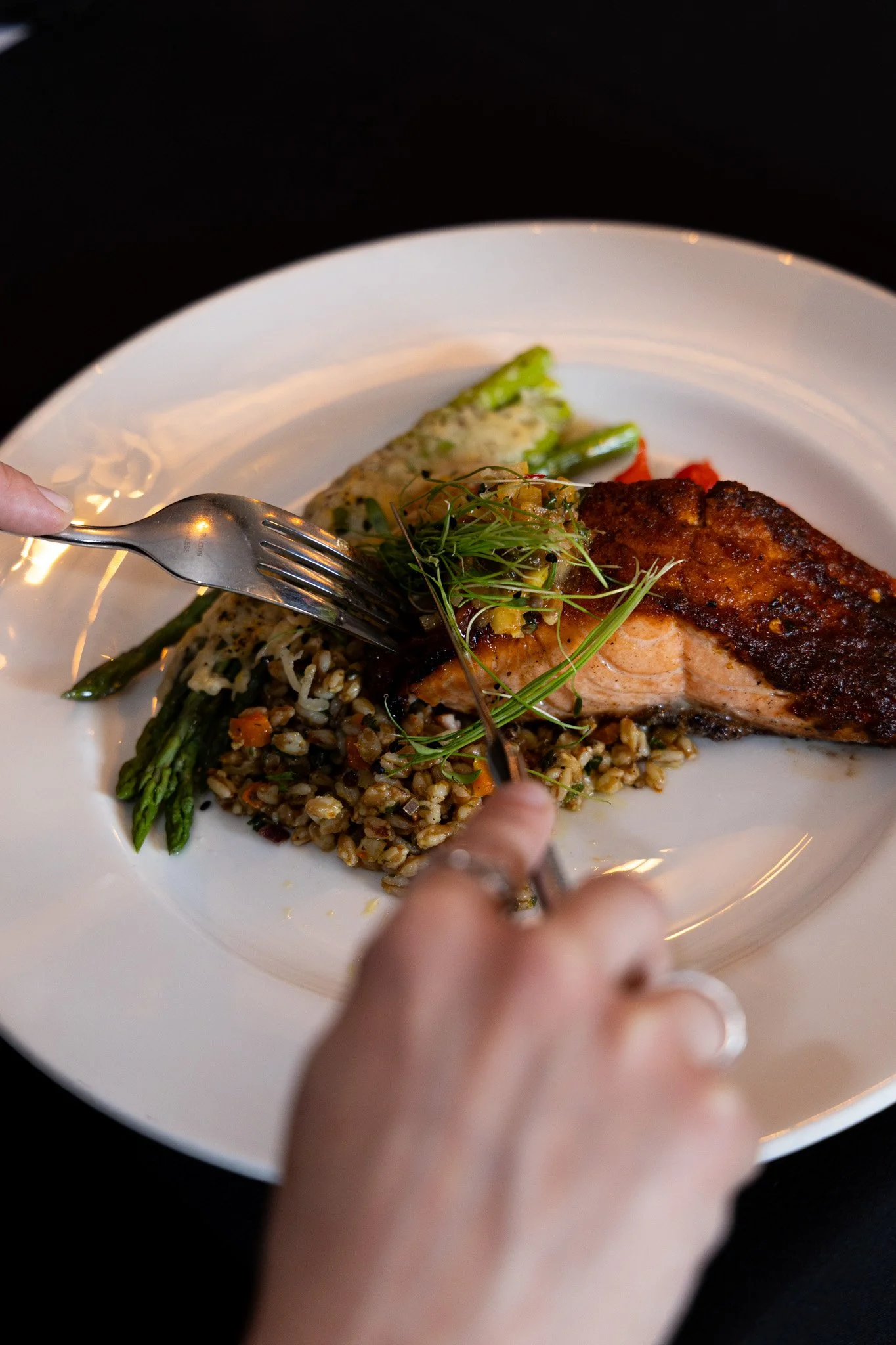 A plate of food with salmon, asparagus, grain, and vegetables, with a hand holding a fork and knife.