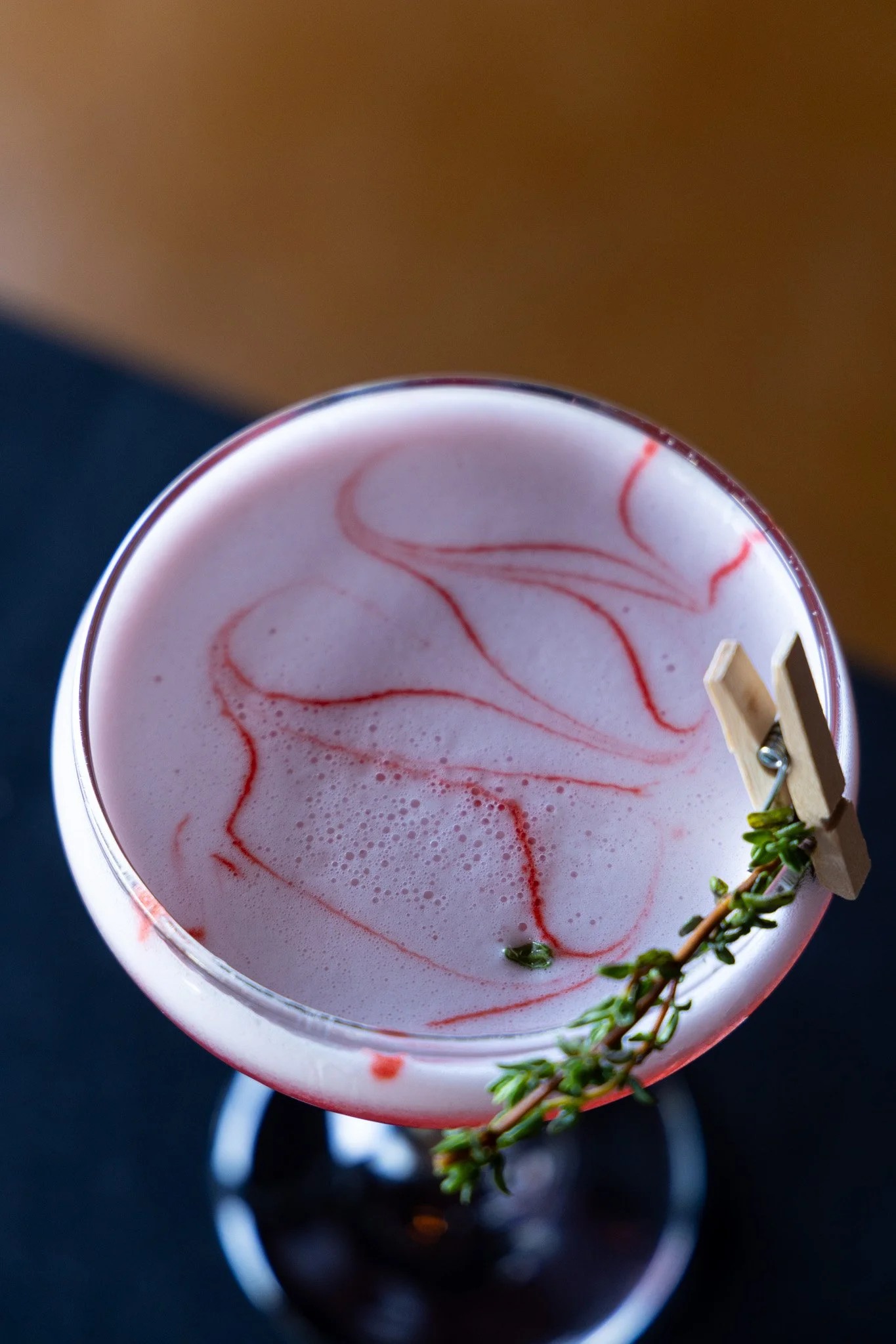 A pink cocktail with red swirl patterns, garnished with a small sprig of thyme, seen from above.