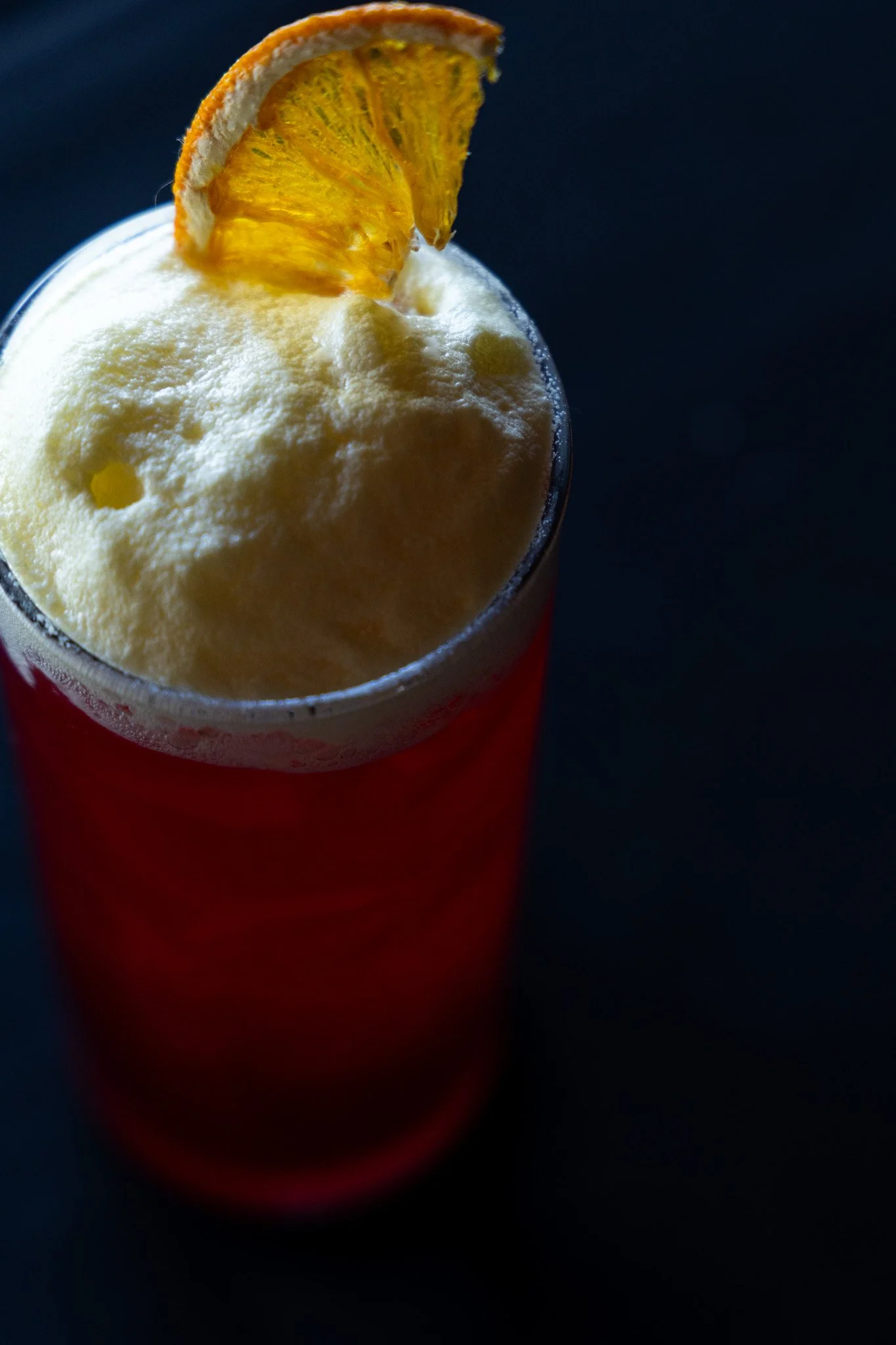 A tall glass of a red cocktail topped with a craft cocktail foam, garnished with a dried orange slice on the rim, against a dark background.