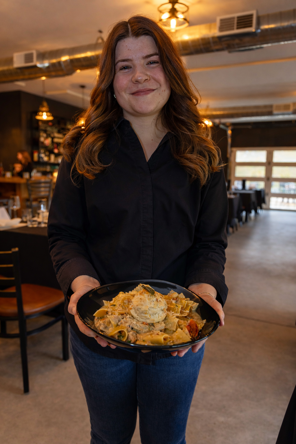 A smiling woman with wavy hair holding a black plate of pasta, inside a restaurant with warm lighting and wooden decor.