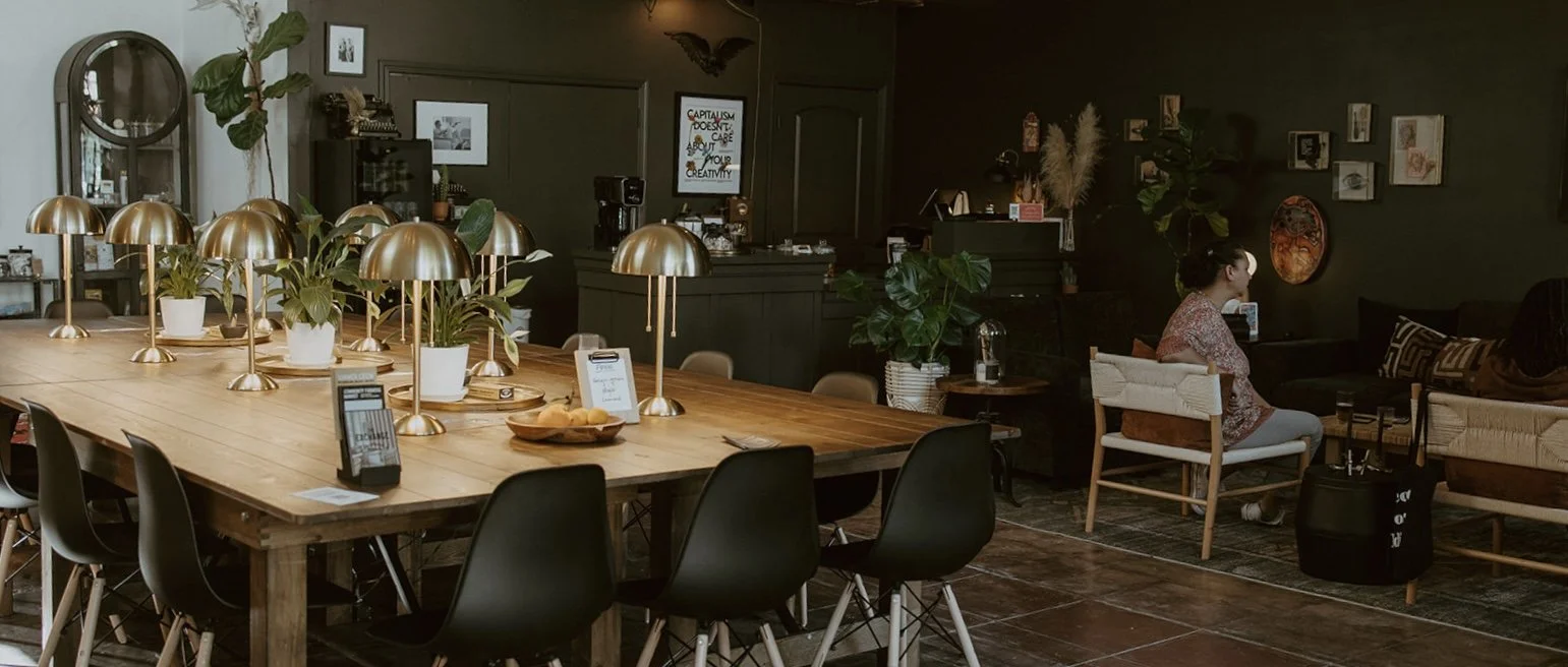 A dining area in a cafe with a large wooden table, multiple brass lamps, and plants. To the right, a woman sits on a white chair at a cozy seating area with couches and artwork on dark-colored walls.