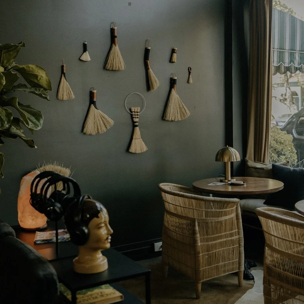 Interior of a cozy restaurant with bamboo decoration on a wall, wicker chairs, a round wooden table with a gold lamp, and a large window with curtains. A mannequin head with headphones is on a side table next to a salt lamp.