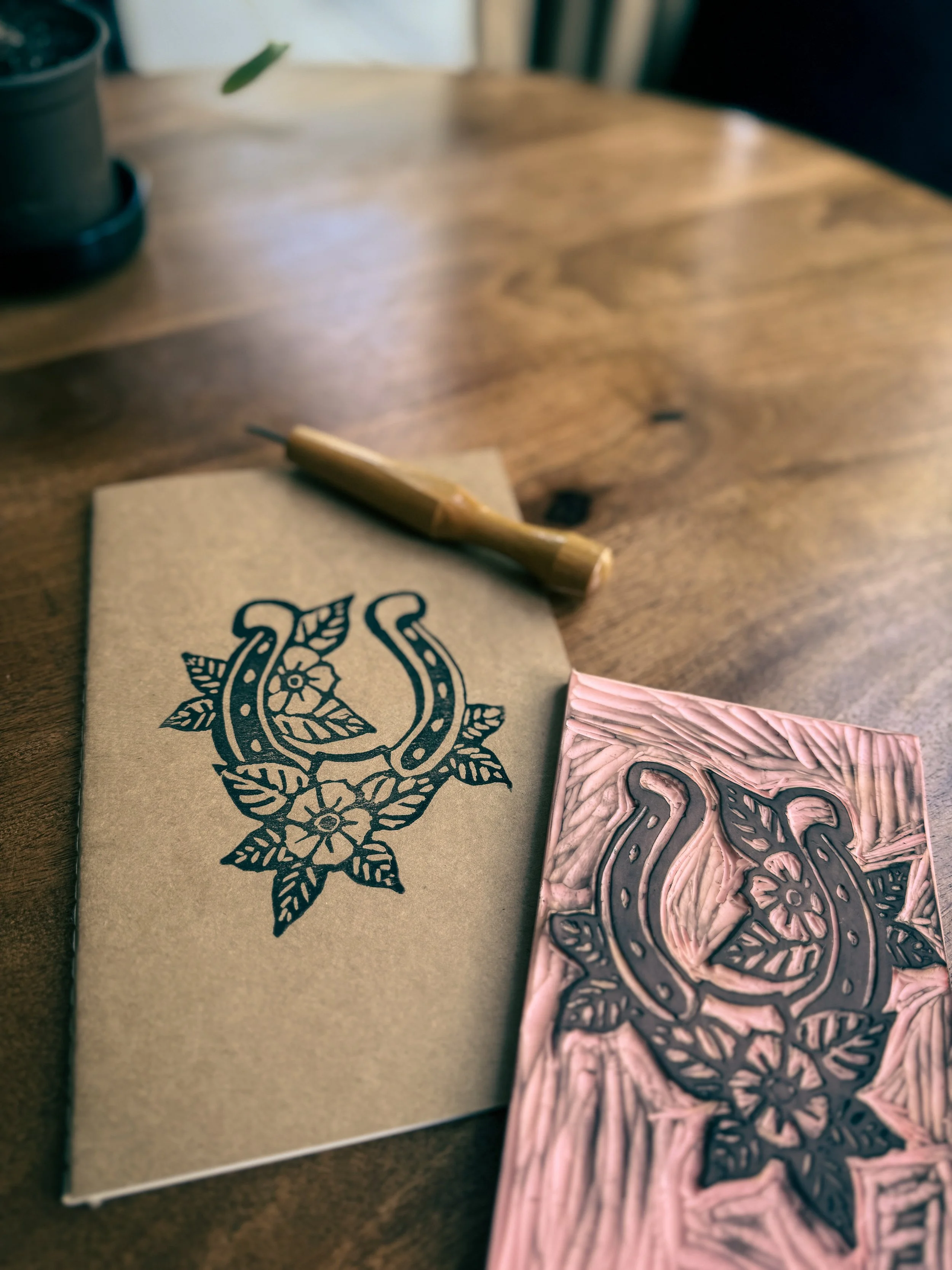 A wooden table with a beige card featuring a black printed floral and horseshoe design, a pink rubber stamp with the same design, and a small wooden-handled stamp pad in the background.