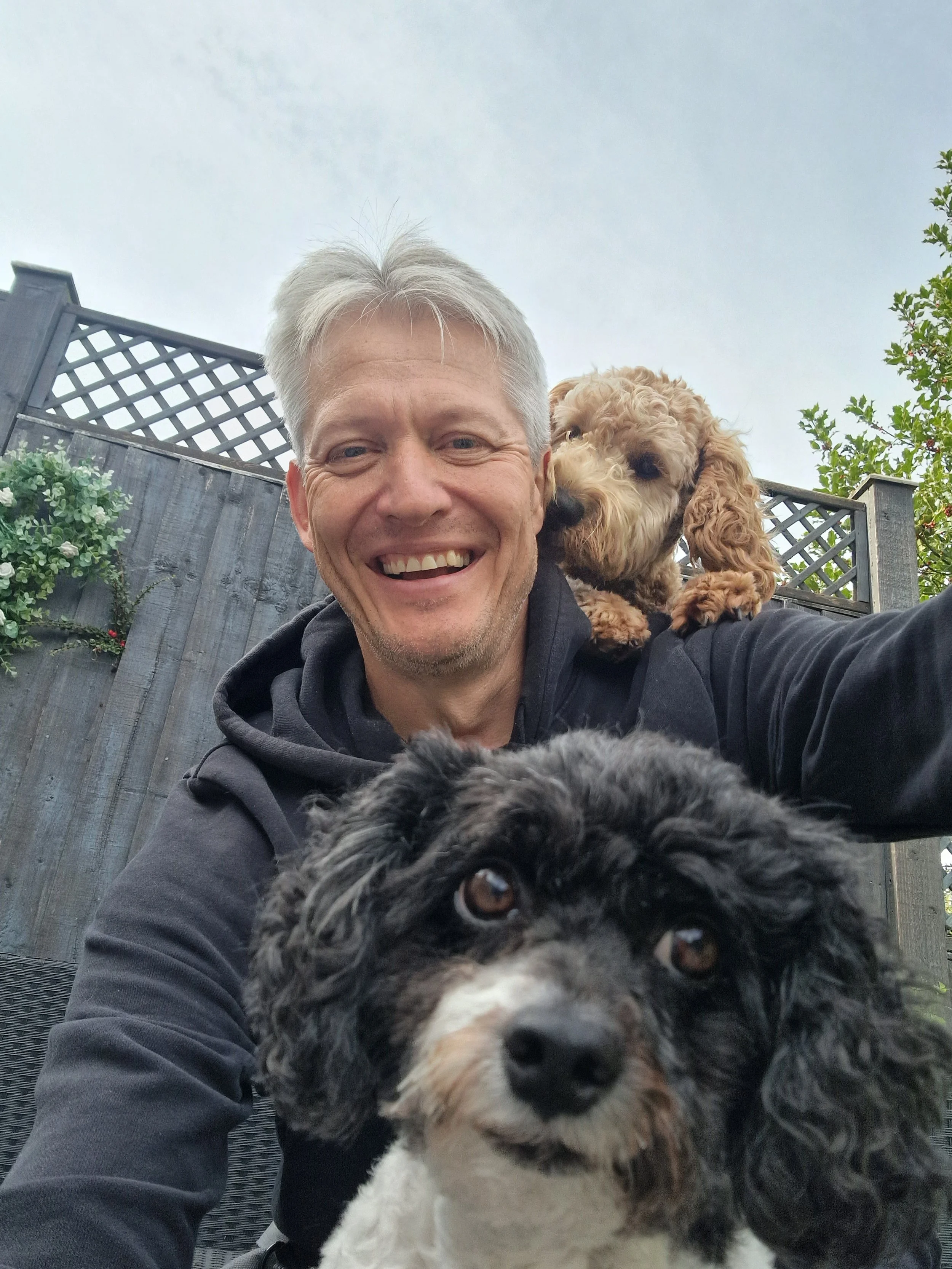 A person smiling with two small dogs outdoors, one black and white dog in the foreground and a brown dog on the person's shoulder. A wooden fence with plants is in the background.