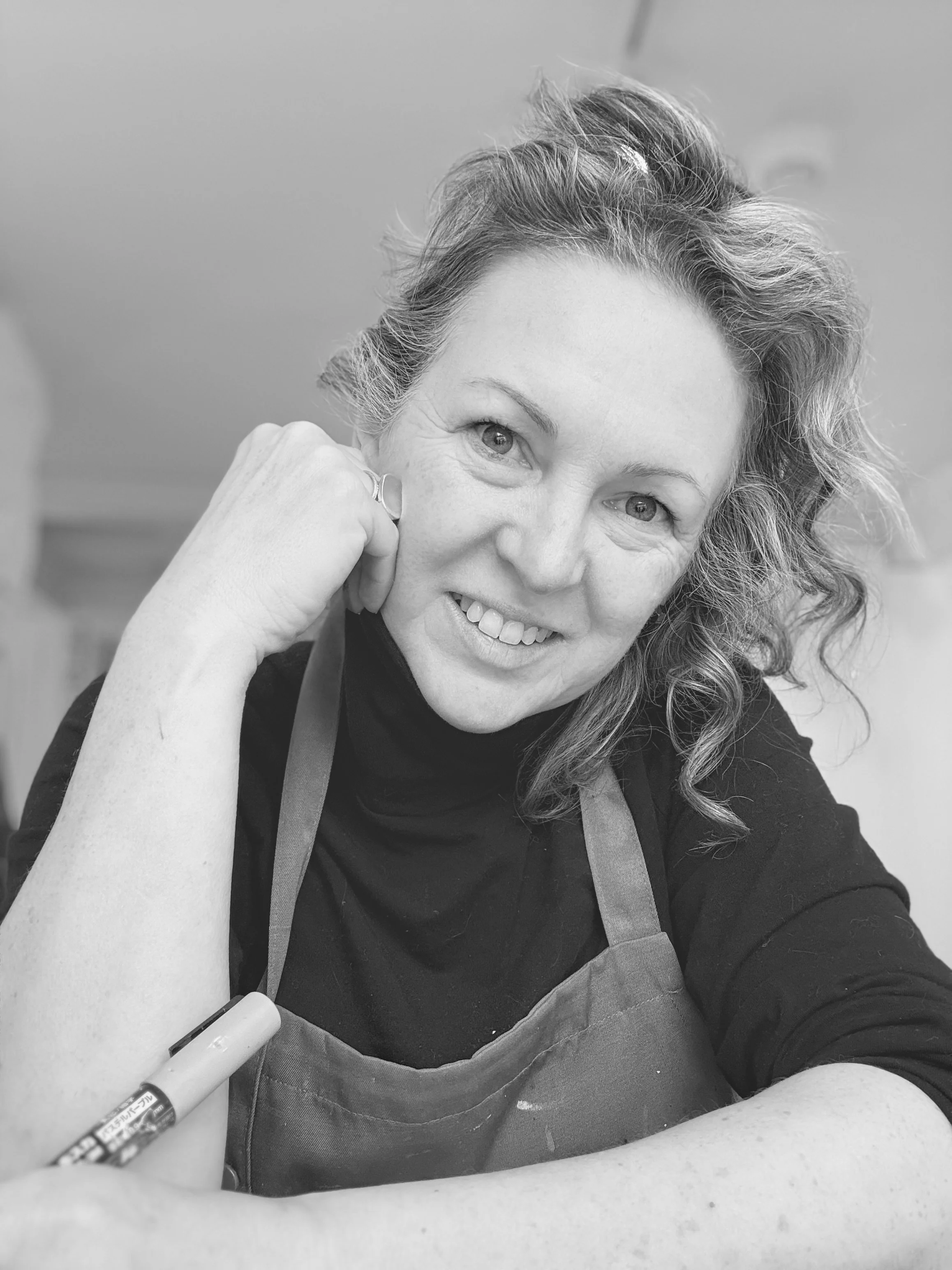 A woman with curly hair smiling at the camera, wearing an apron, with her arm resting on a table, holding a marker in her hand.