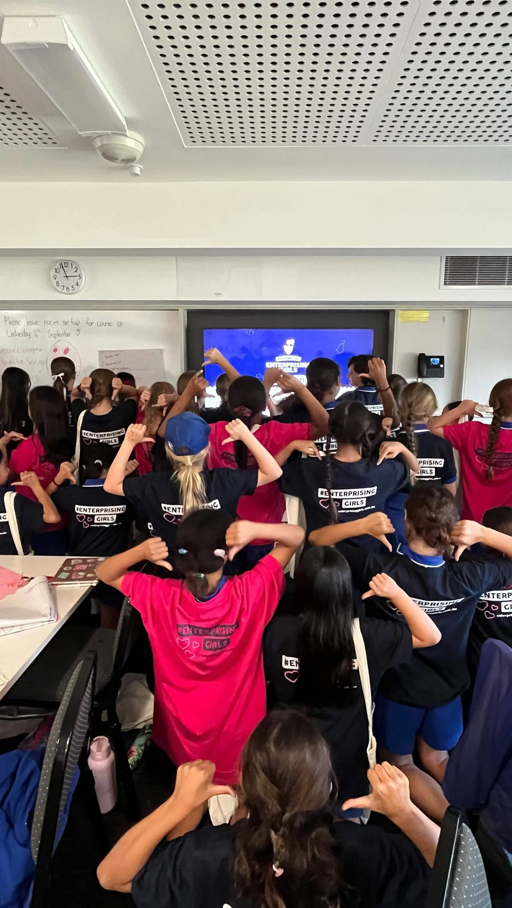 Group of children in a classroom facing a screen, wearing shirts with "Enterprising Girls" on the back, pointing to the text. The screen displays a presentation. Classroom setup with chairs and a clock visible.