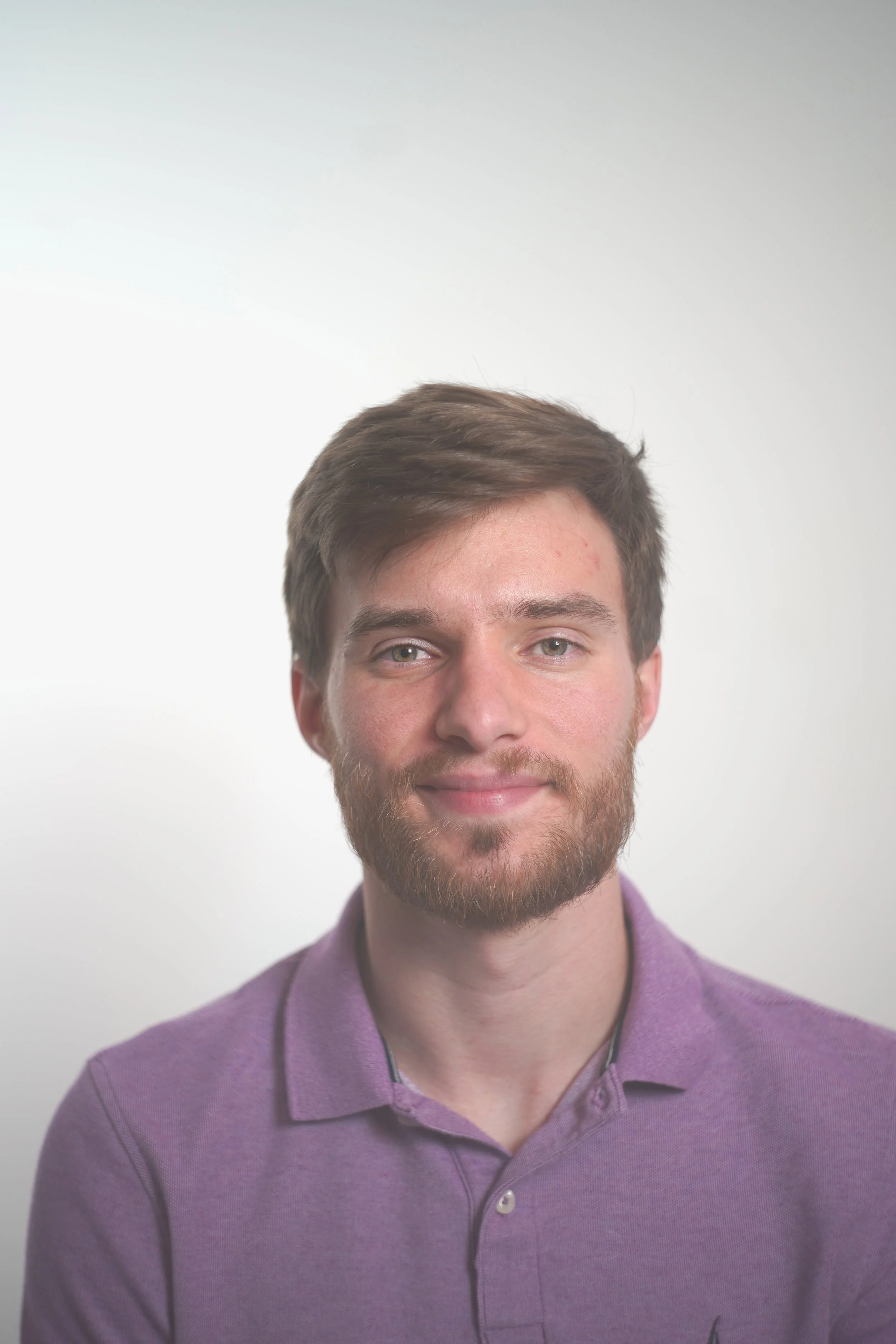 Headshot of a young man with brown hair, beard, light-colored eyes, wearing a purple collared shirt, smiling softly, plain light background.