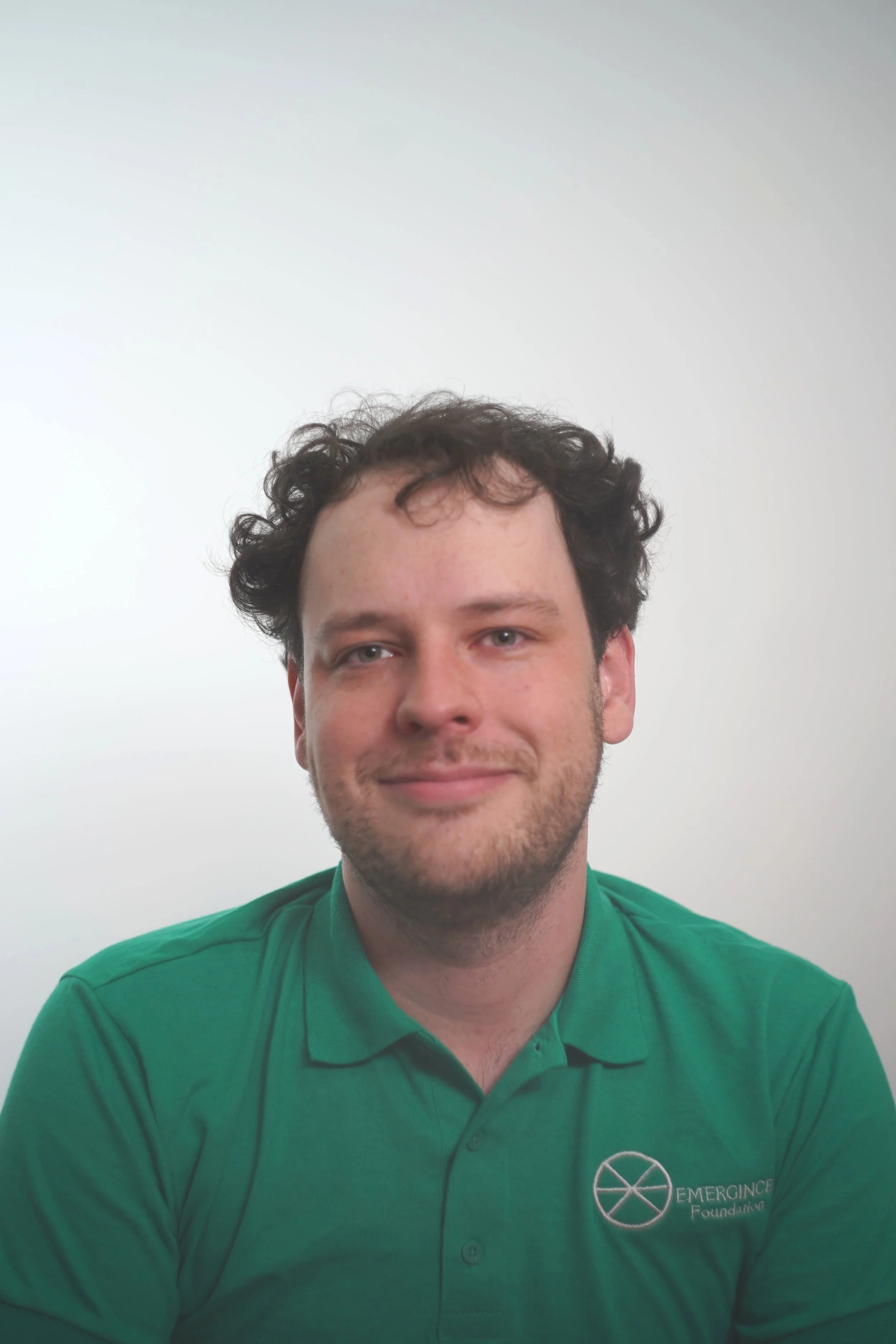 A young man with curly brown hair wearing a green polo shirt with a logo that says 'Emerging Foundation' on a plain white background.