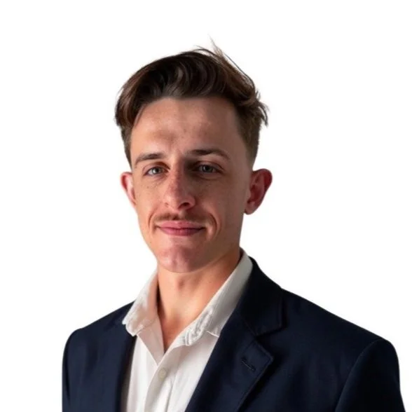 Portrait of a young man with light skin, short brown hair, wearing a navy blazer and white shirt, standing against a plain white background.