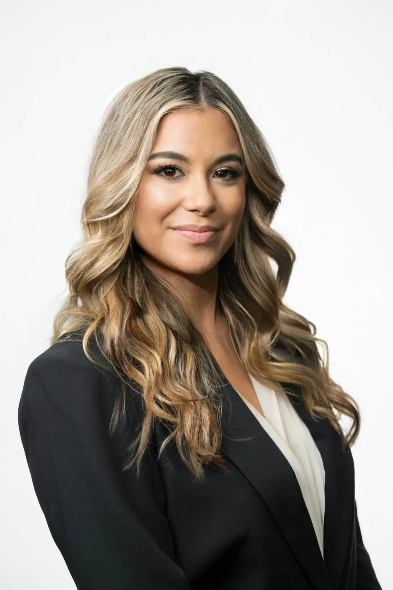 Professional woman with long wavy blonde hair in a black blazer and white blouse, smiling confidently against a plain white background.