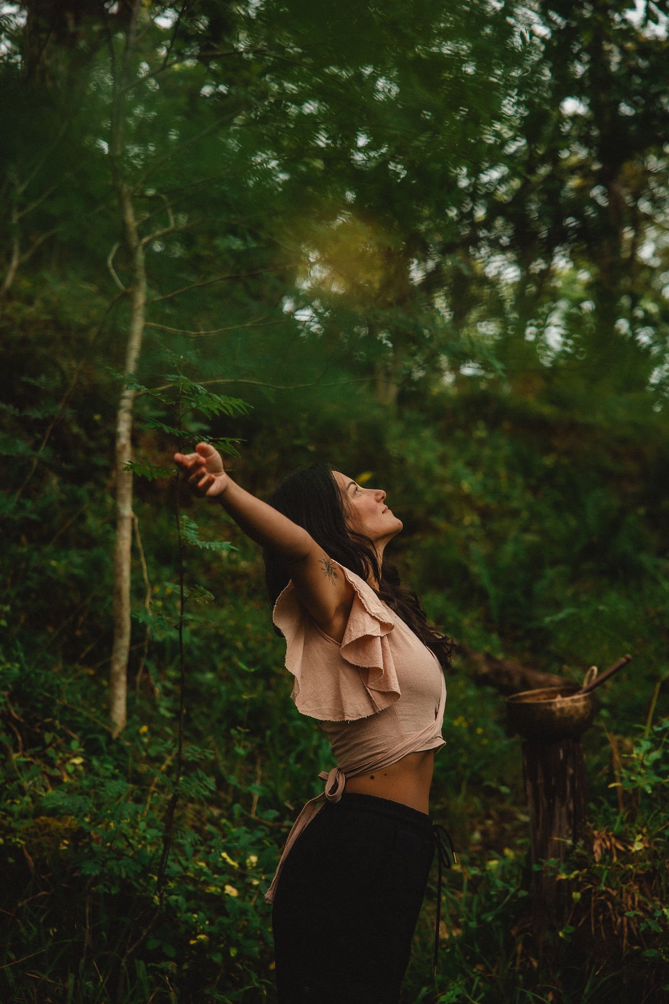 Lara Wehbe somatic therapy Glasgow in a lush green forest with eyes closed and arms outstretched, reaching towards the sky.