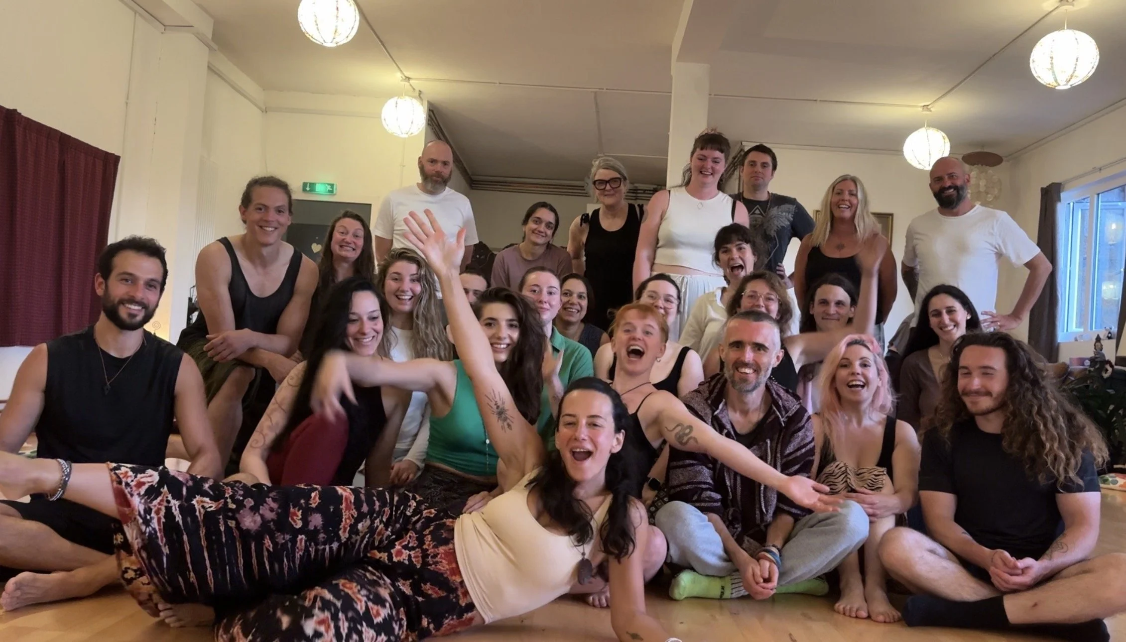 A large group of people smiling and posing together indoors. They are sitting or standing in a cozy room with warm lighting, curtains, and hanging lamps.