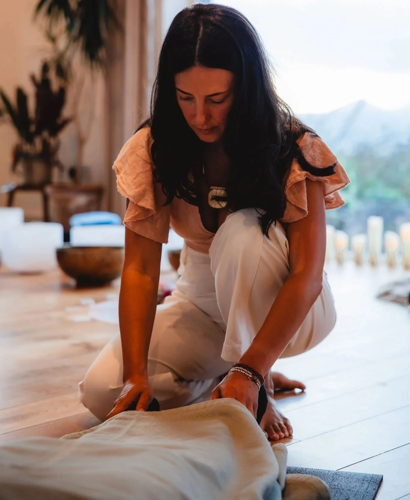 A woman with long dark hair, wearing a peach-colored top and beige pants, kneels on the floor and appears to be creating a sand mandala or engaging in a similar meditative activity. There are bowls and candles in the background, indicating a peaceful, spiritual setting.