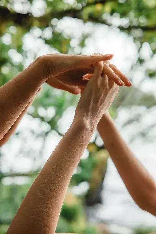 Close-up of multiple hands with intertwined fingers, raised against a backdrop of blurred green leaves and sky.