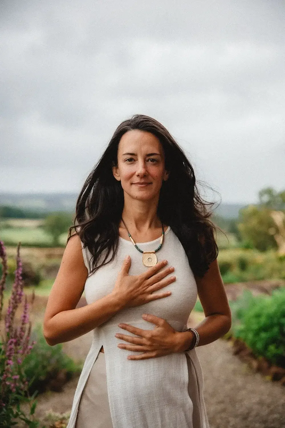 A woman with long dark hair standing outdoors on a cloudy day, holding her hand over her heart and stomach, wearing a sleeveless white dress and necklaces, with green farmland and purple flowers in the background.