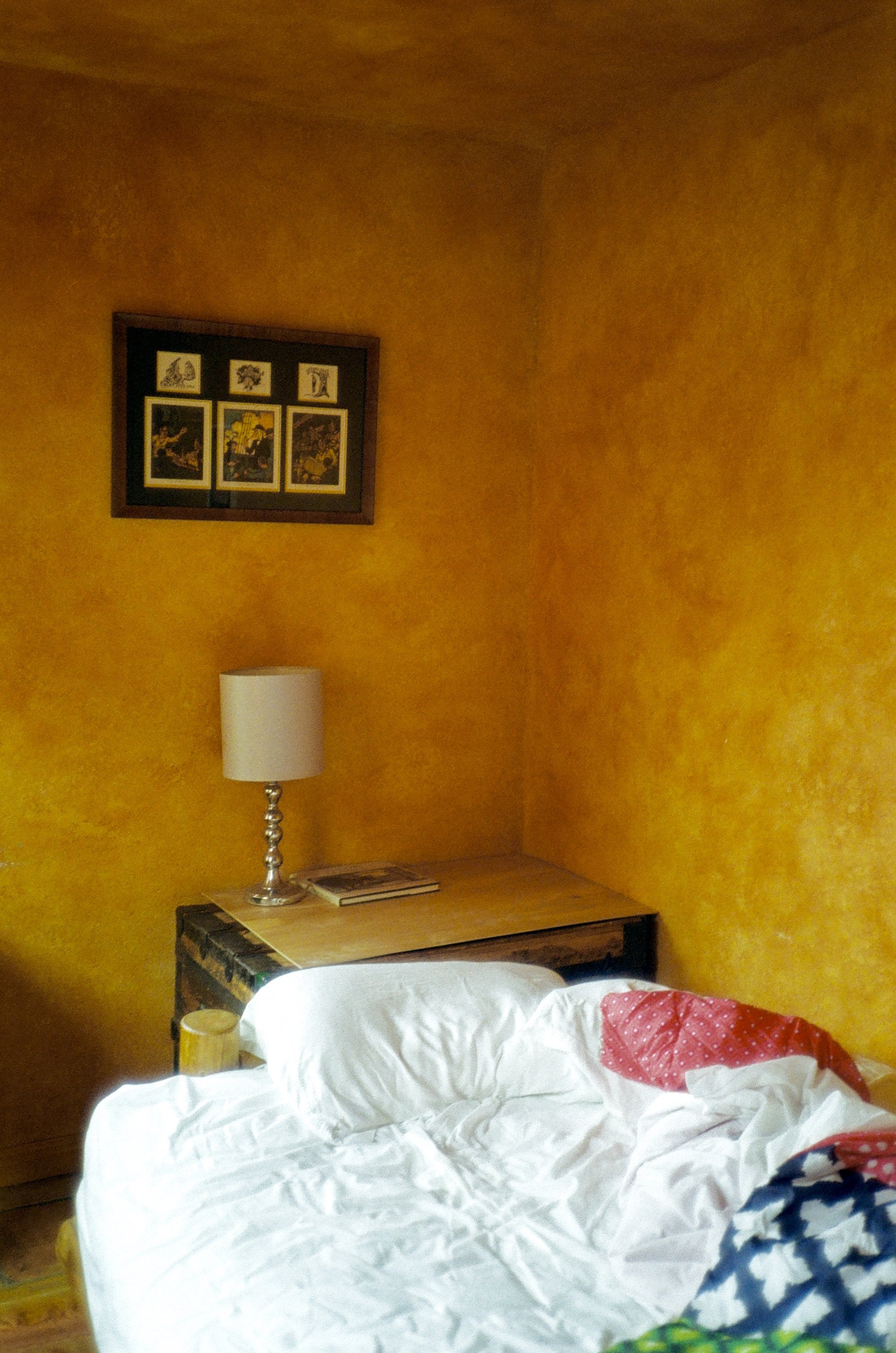 Bedroom with yellow walls, a lamp on a wooden table, framed pictures, and an unmade bed with mixed-pattern bedding.