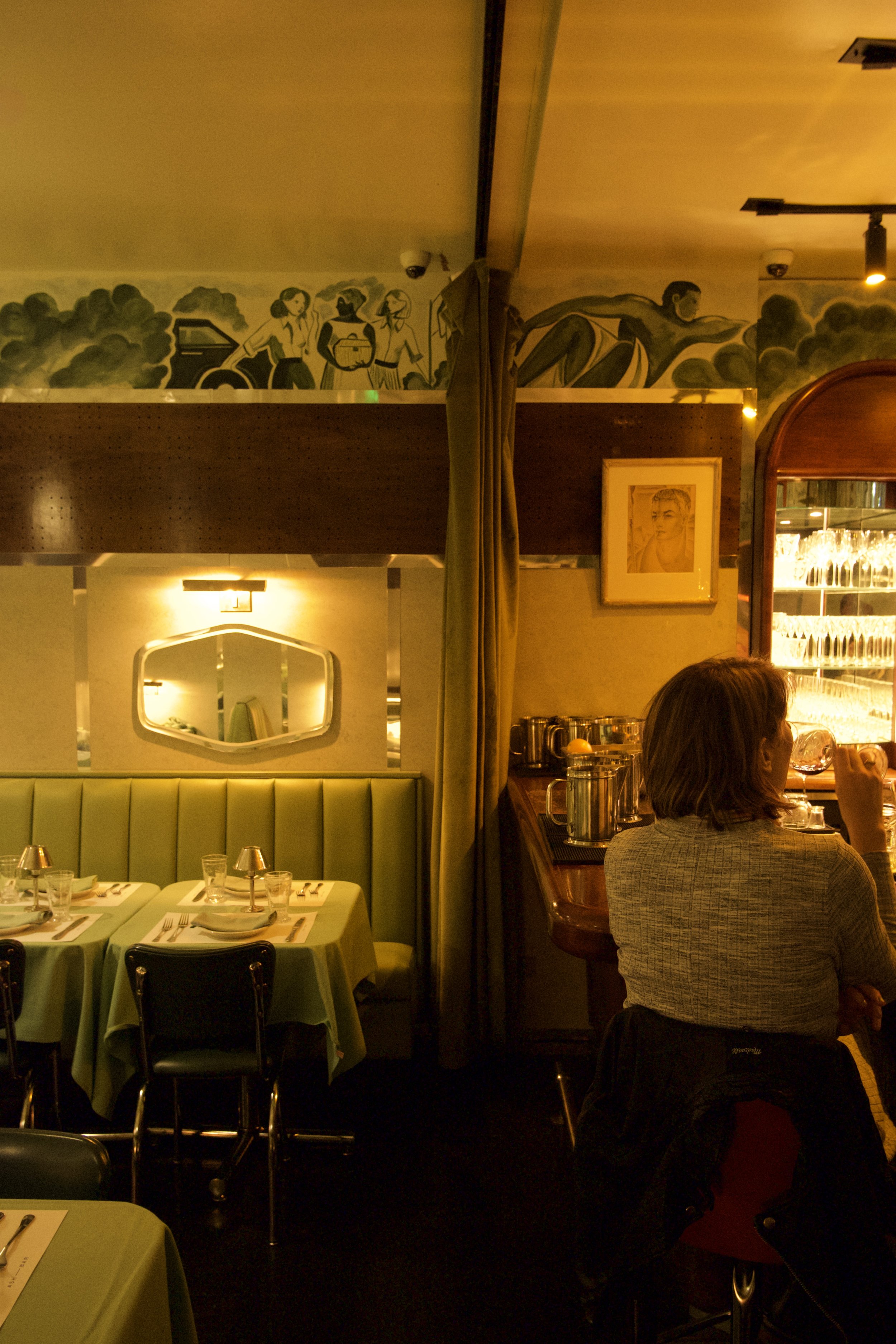 interior of a cozy restaurant with green booth seating, set tables, a mural of people, and a woman sitting at a bar