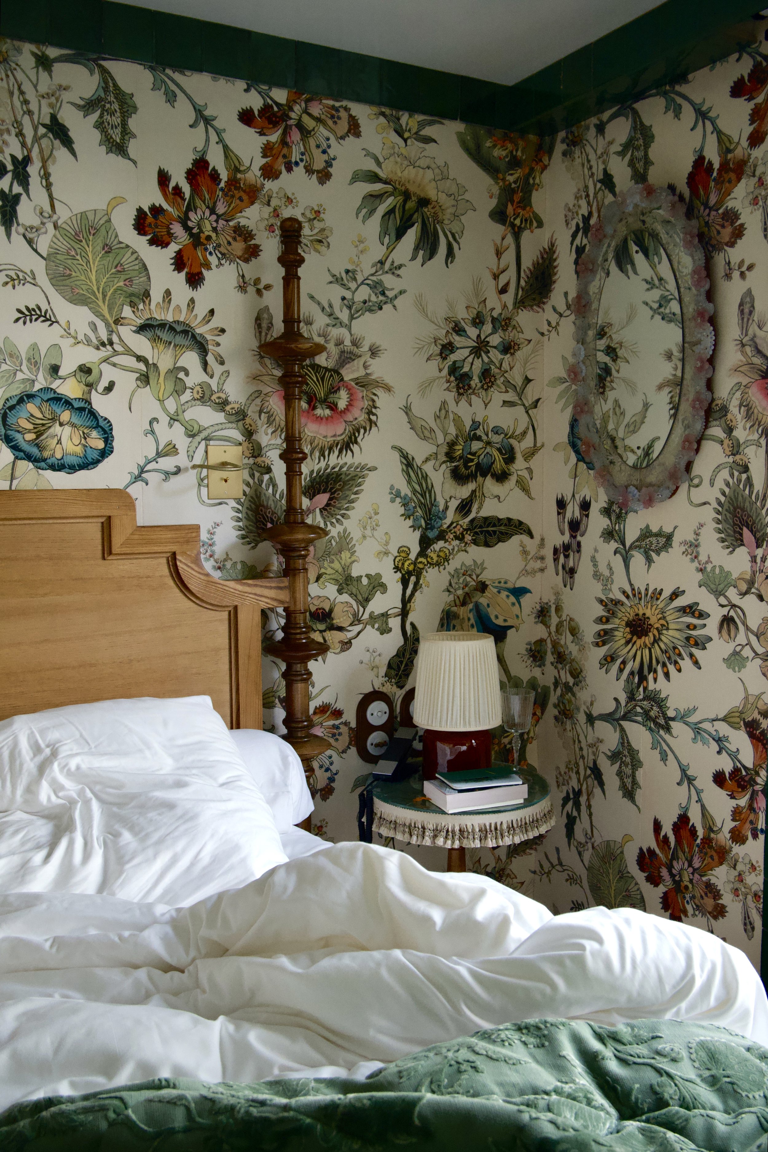 Cozy bedroom with floral wallpaper, wooden bedpost, and white bedding. A nightstand holds a lamp, books, and decorative items. An ornate mirror is on the wall.