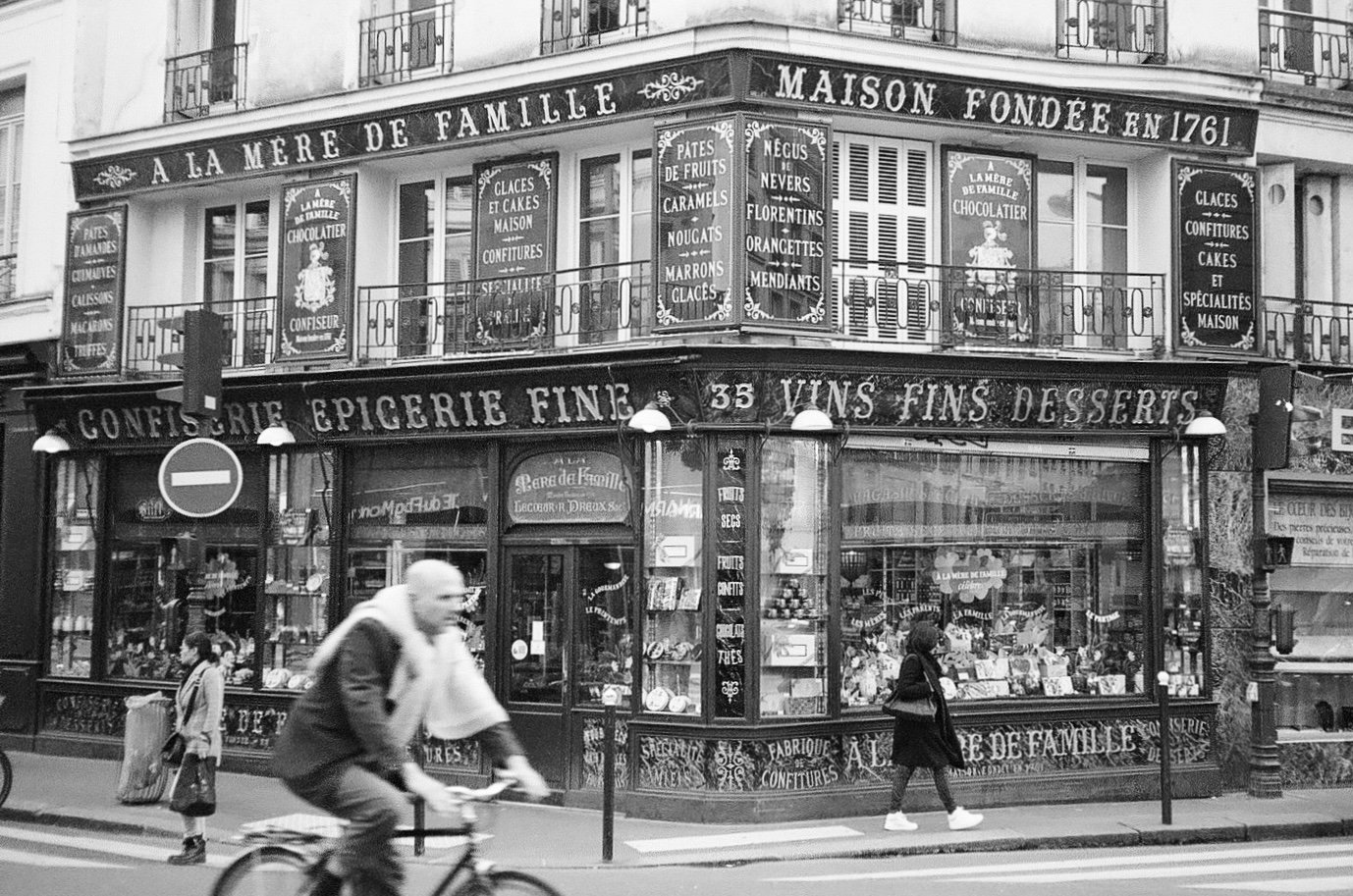 Vintage Parisian shopfront "A La Mère de Famille" with a cyclist and pedestrian in foreground, black and white photograph.