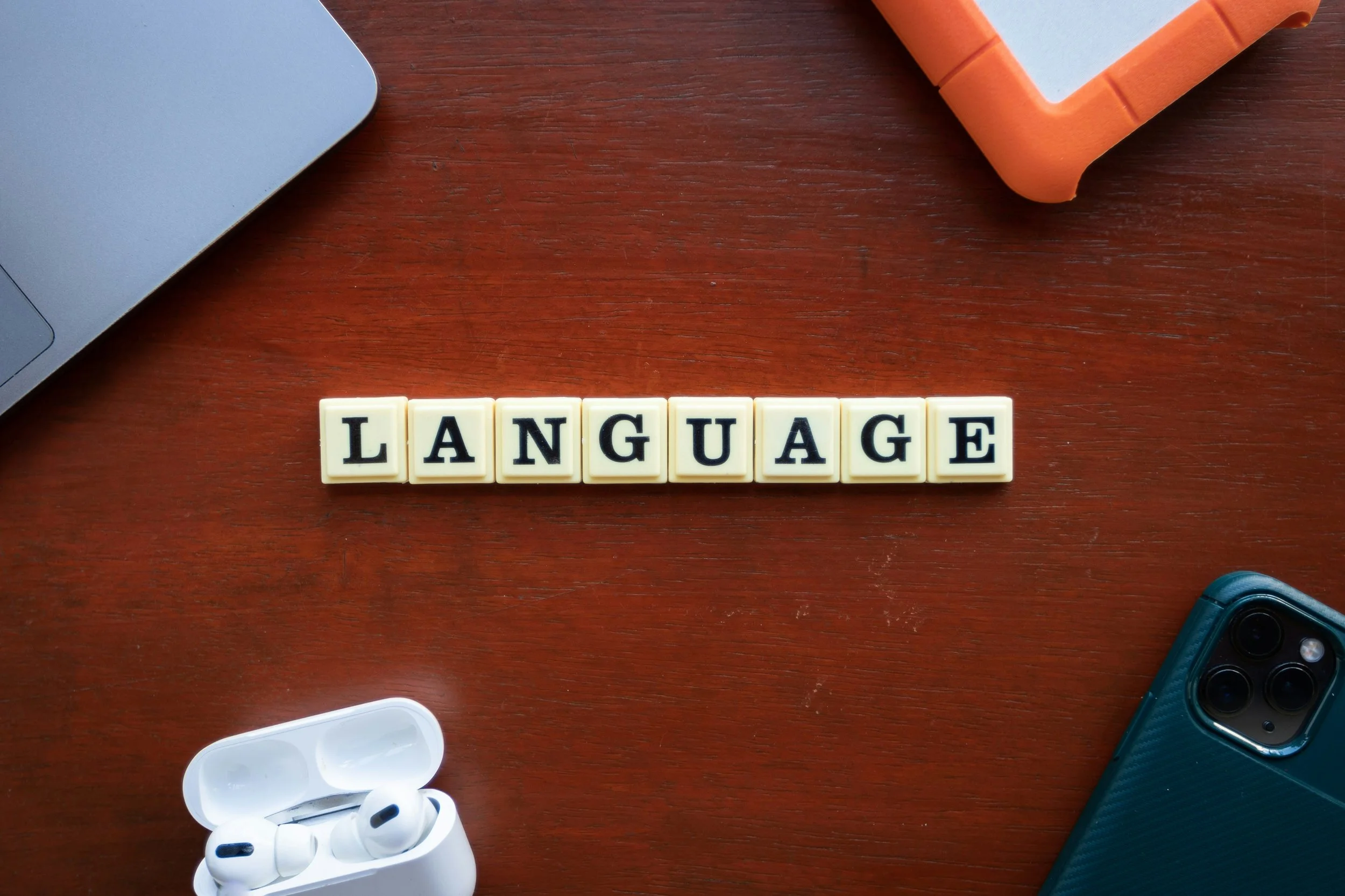 A wooden surface with white Scrabble tiles spelling 'LANGUAGE' at the center, a gray laptop on the top left, orange and white containers on the top right, a blue phone on the bottom right, and white wireless earphones with a charger on the bottom left.