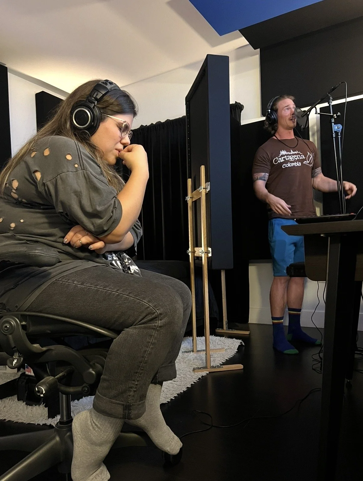  Brooke Markham wearing headphones and glasses, sitting on a rolling chair with crossed arms and a thoughtful expression, in a recording studio. Aaron Cavette is performing.