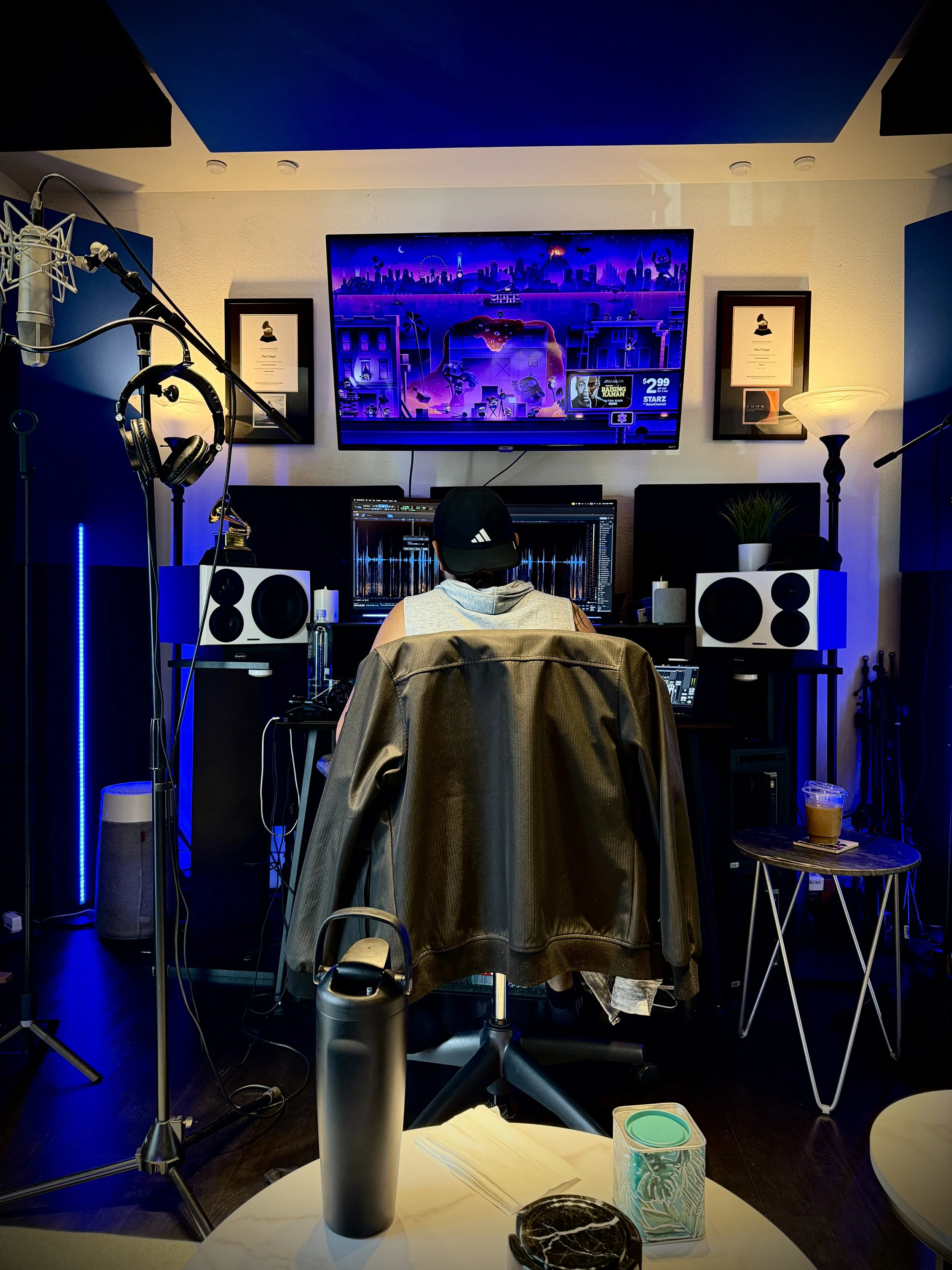 Photo of Raul Vega in studio with music editing software on the computer screen, surrounded by studio equipment, and a cityscape on the TV screen in the background.