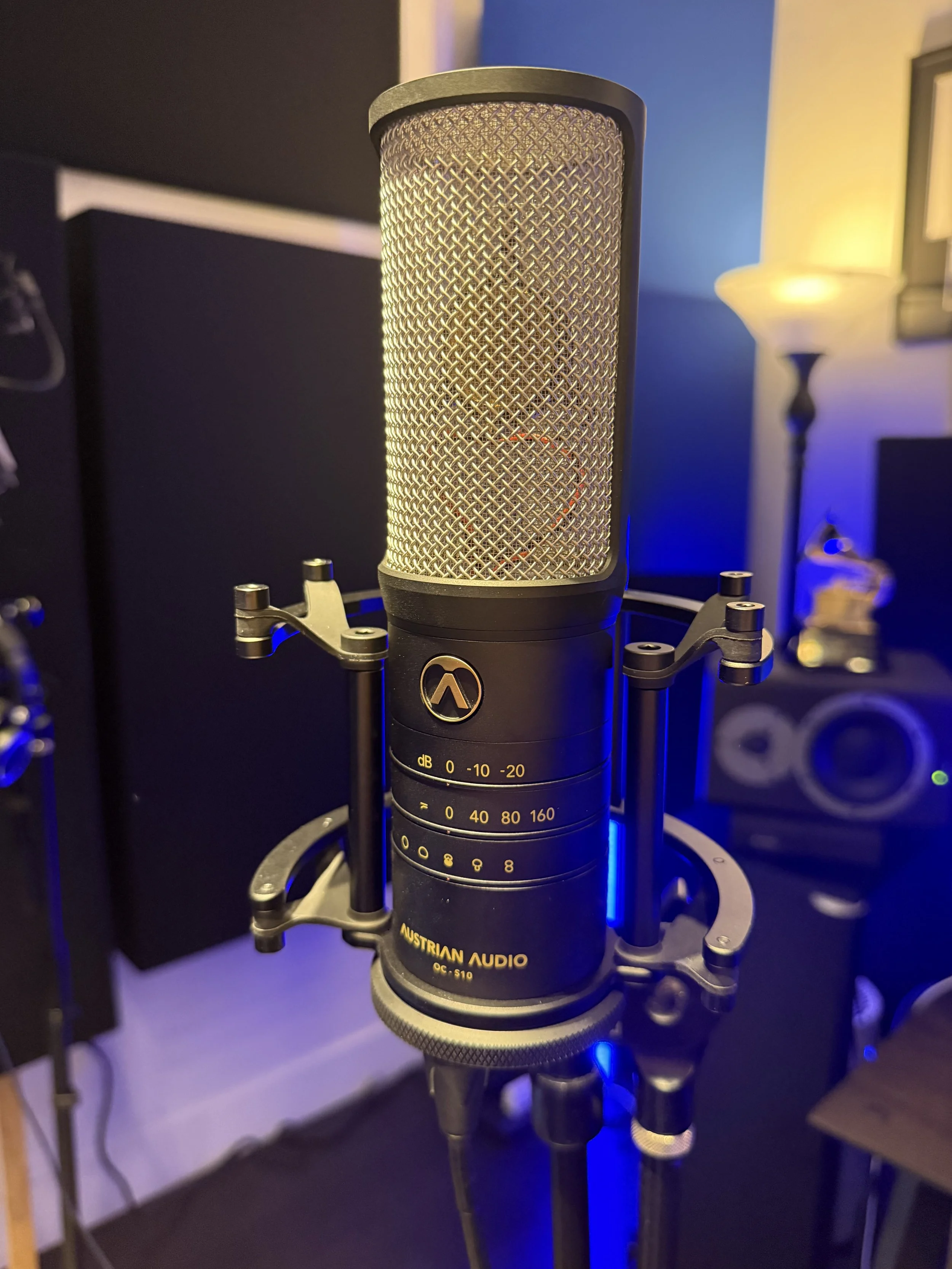 Close-up of a large-diaphragm condenser microphone mounted on a stand, labeled Austrian Audio, OC S10, in a recording studio with dark walls, a blue-lit lamp, and studio equipment in the background.