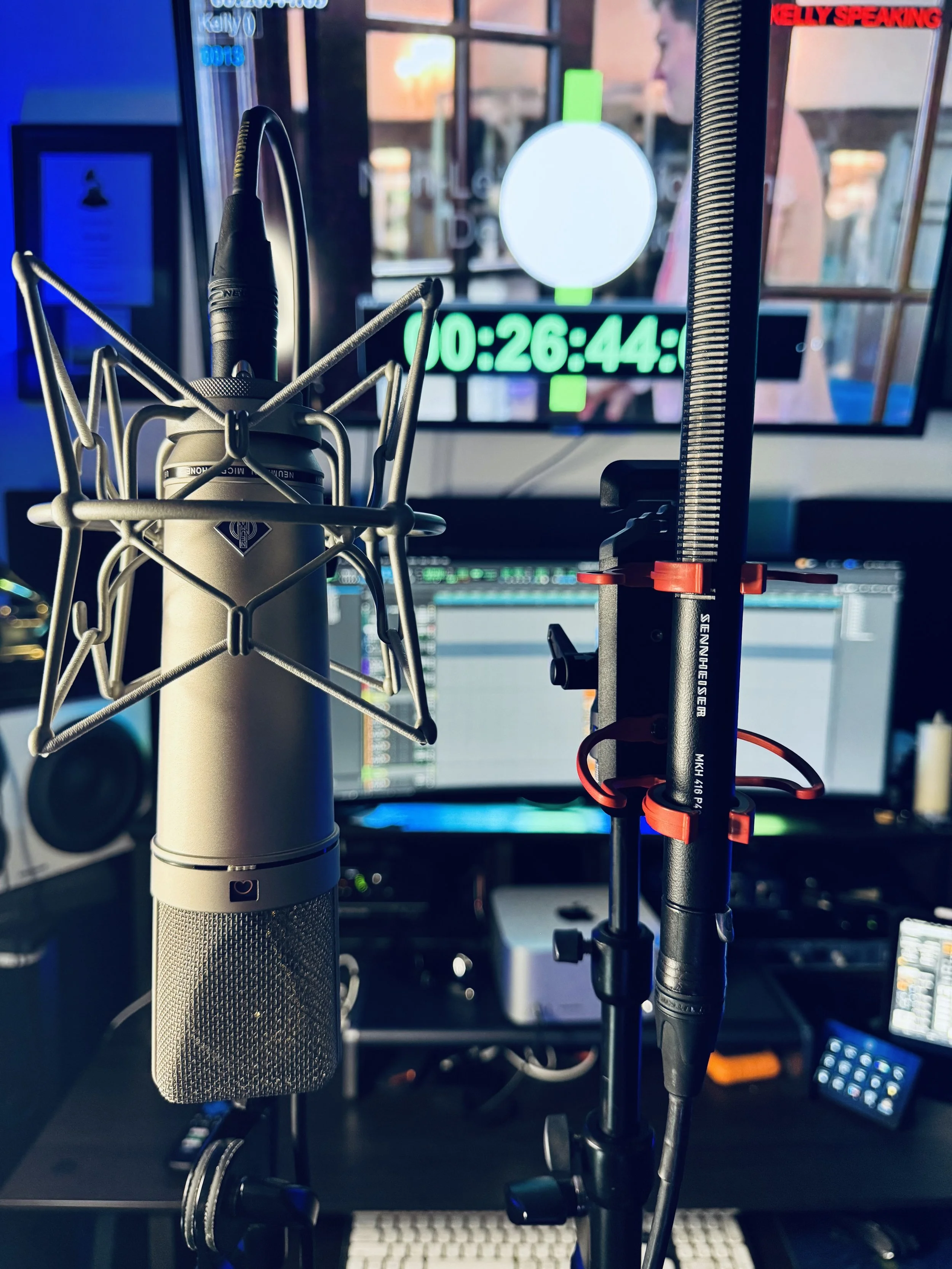 Microphone setup with shock mount and pop filter in front of a computer monitor displaying a timer and a weather report, with a person blurred in the background.