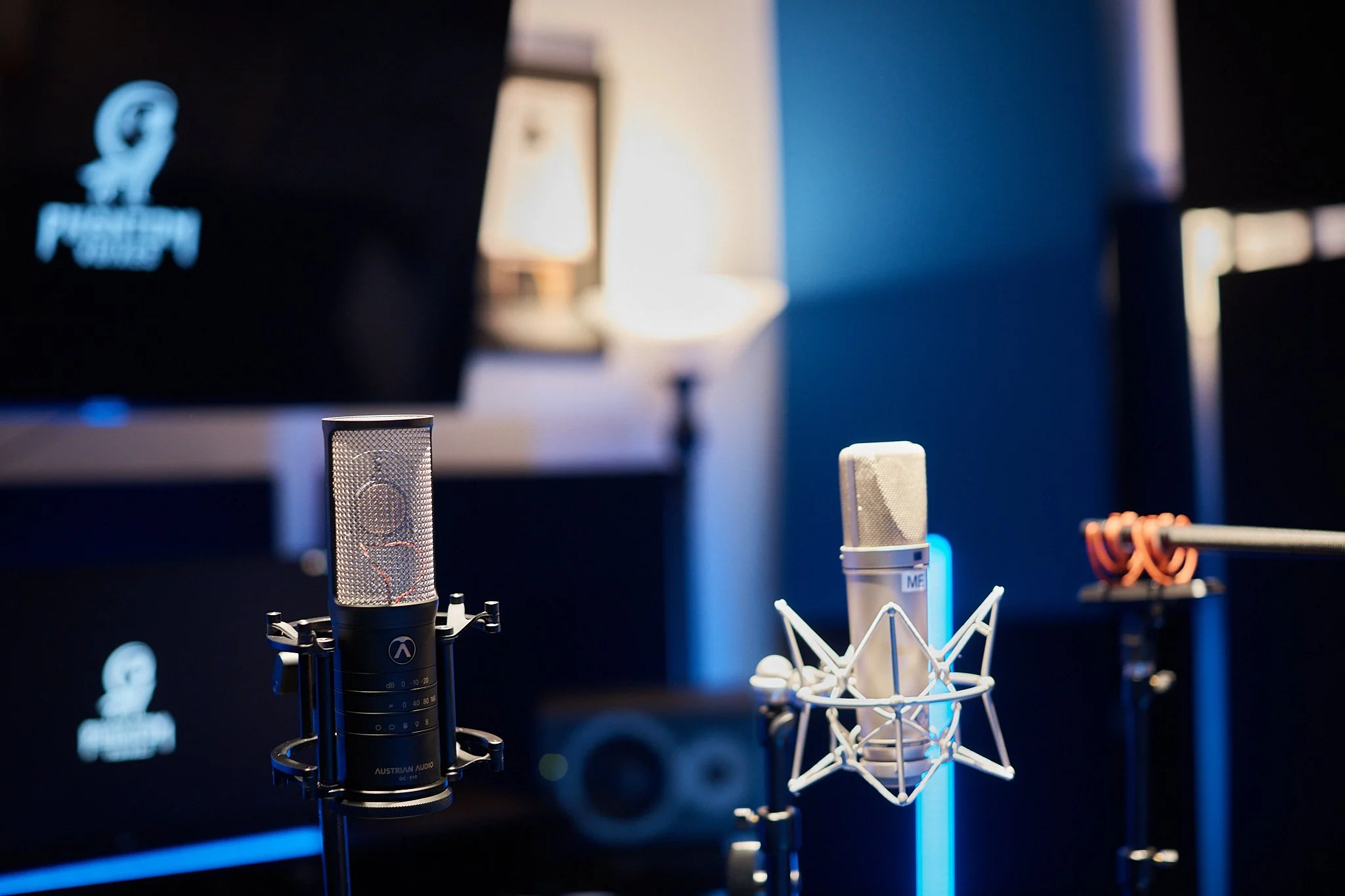 Close-up of professional microphones U87Ai, OC-S10 and MKH416 on adjustable stands in a recording studio with a dimly lit background, including speakers and acoustic panels.
