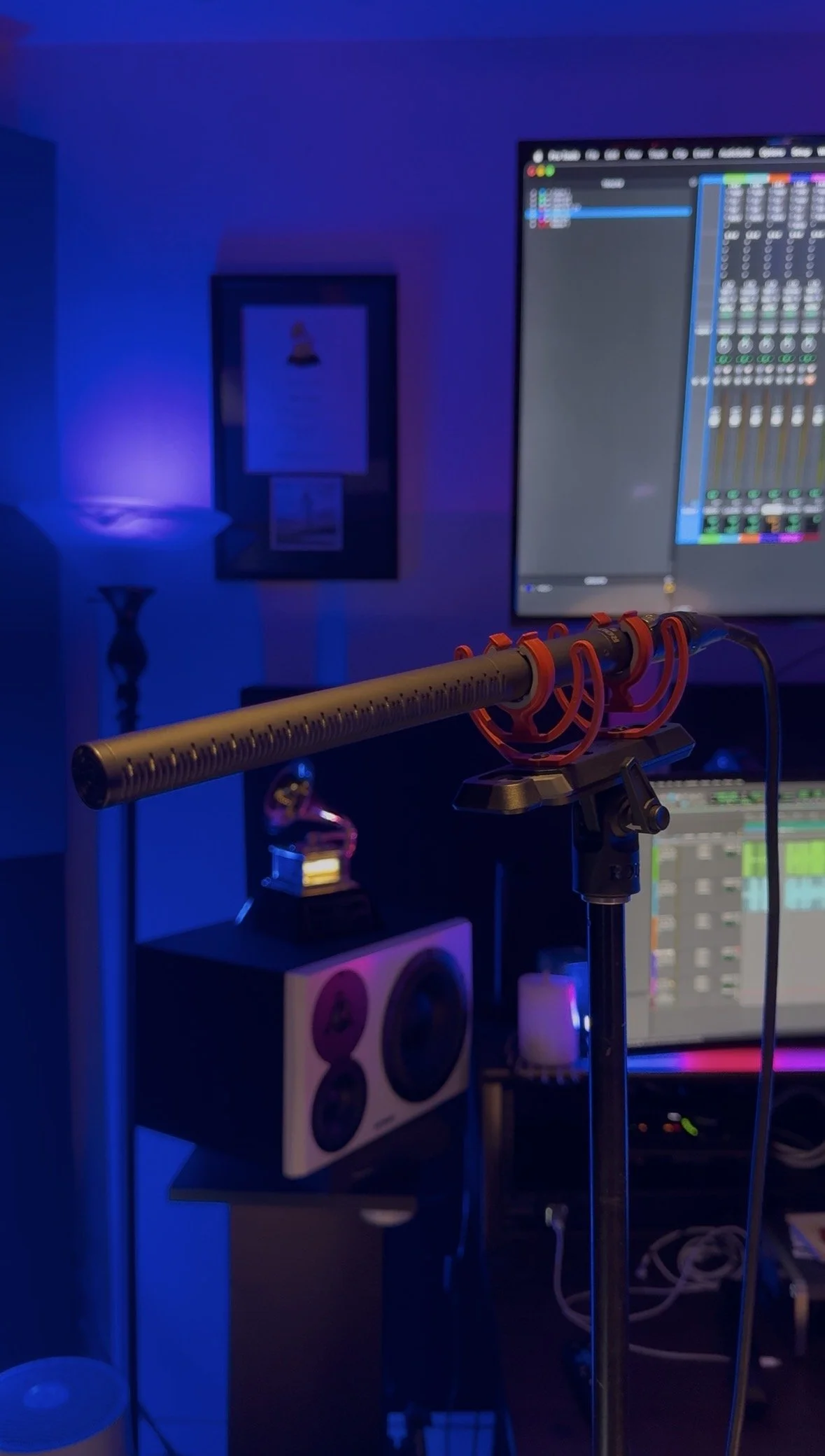 A DJ recording microphone setup in a room with blue ambient lighting, a computer monitor displaying audio editing software, and a wall with framed artwork.