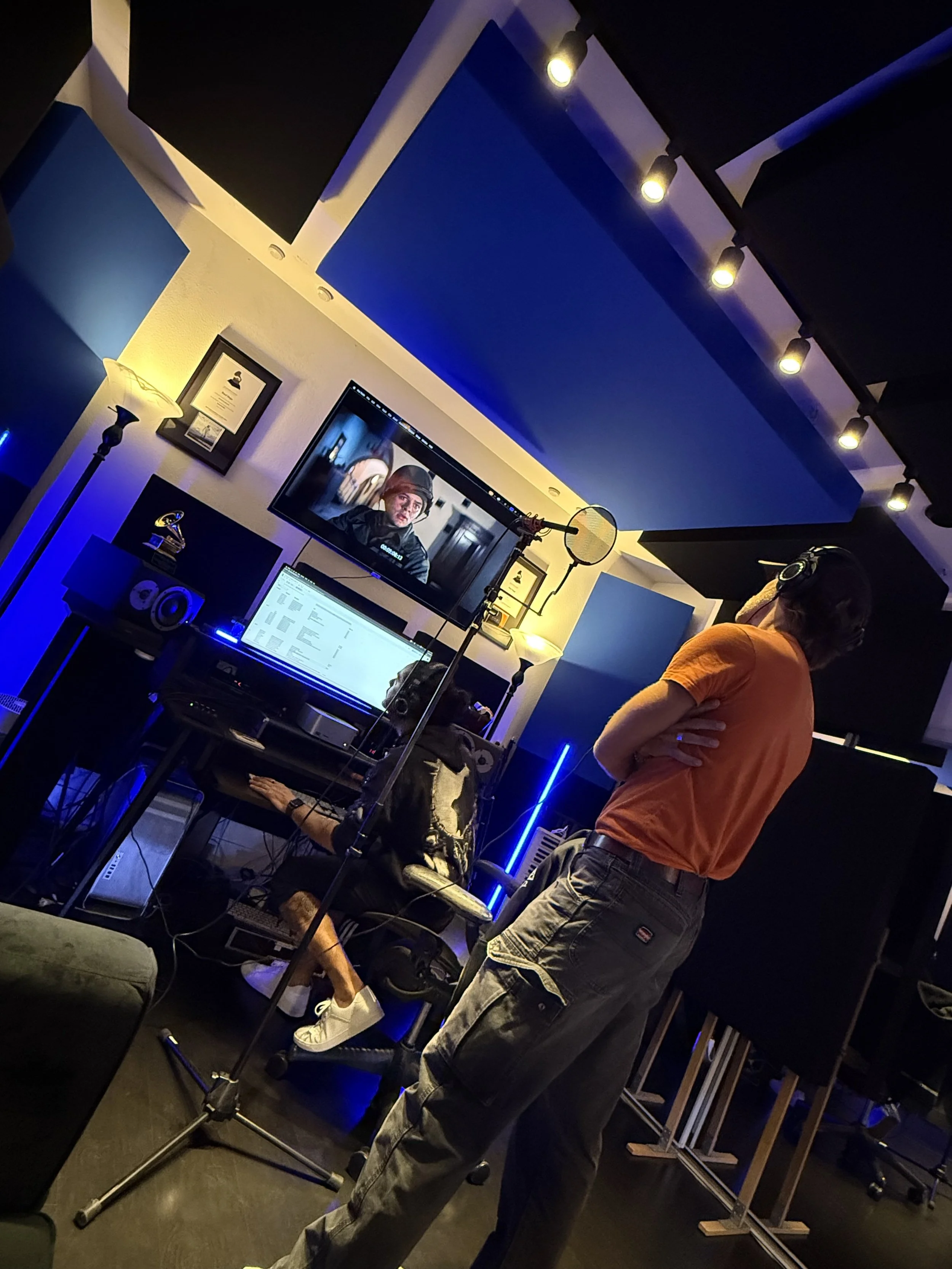 A music recording studio with a person standing in front of a microphone, wearing headphones and an orange shirt, observing a woman seated at a desk with a laptop, there are monitors, speakers, and framed certificates on the wall, with blue and yello