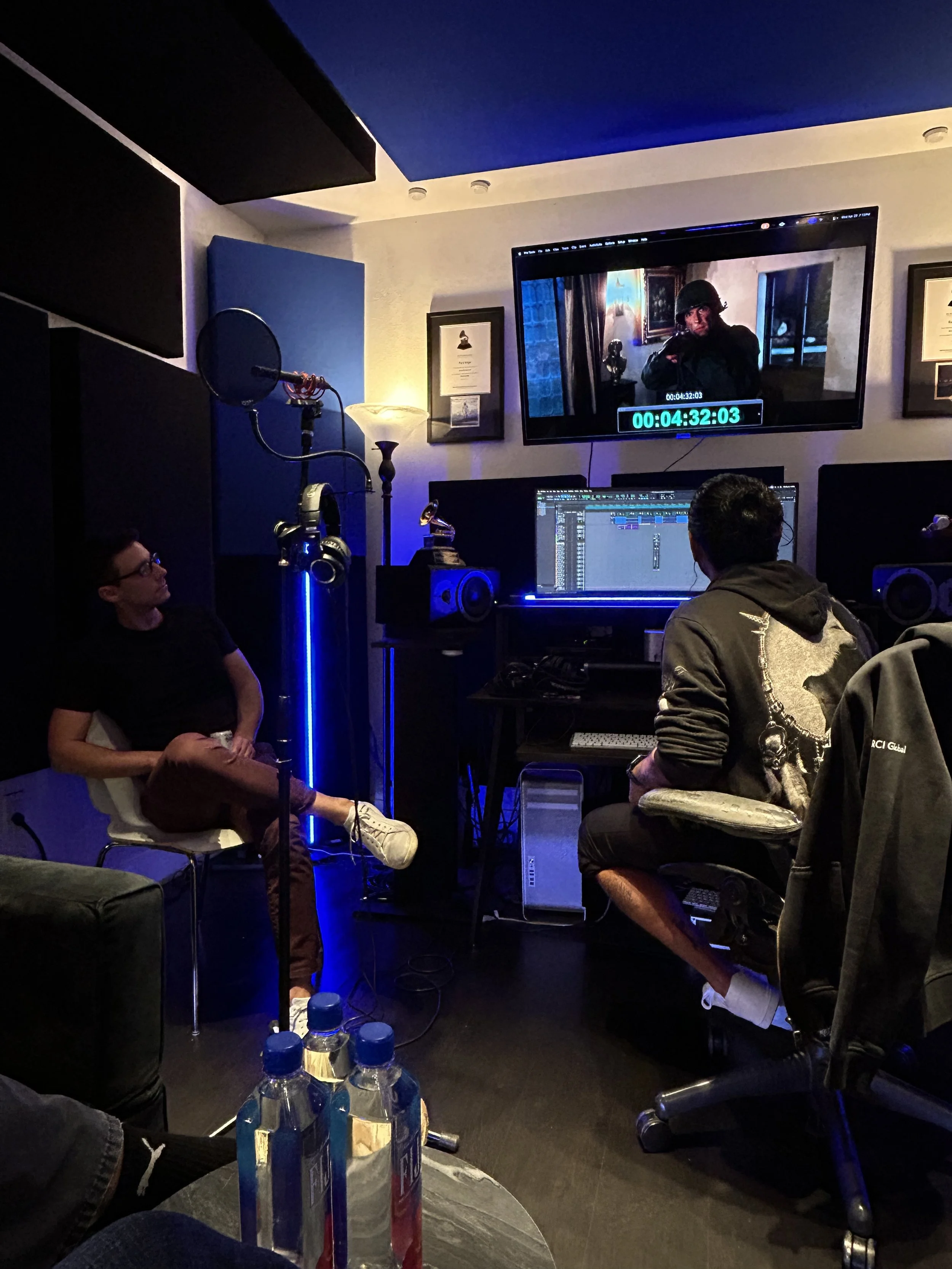 Recording studio with two people working at a computer, a woman sitting on a chair, and a man seated at a desk, with studio equipment and a large monitor displaying a video of a soldier in military gear.