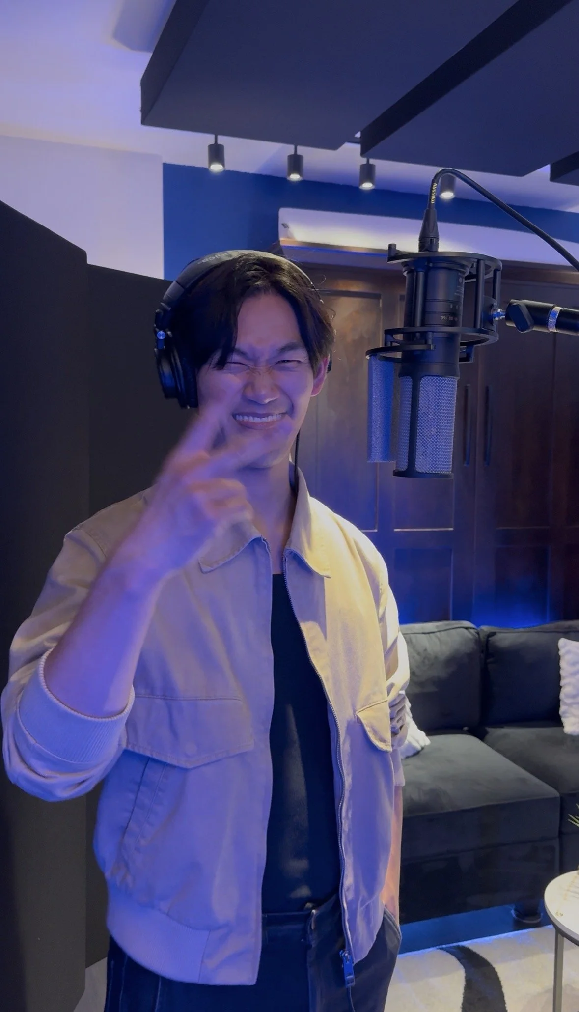 Daniel J Kim in a beige jacket and black shirt, wearing headphones, smiling and pointing with his finger near a professional microphone in a modern recording studio.