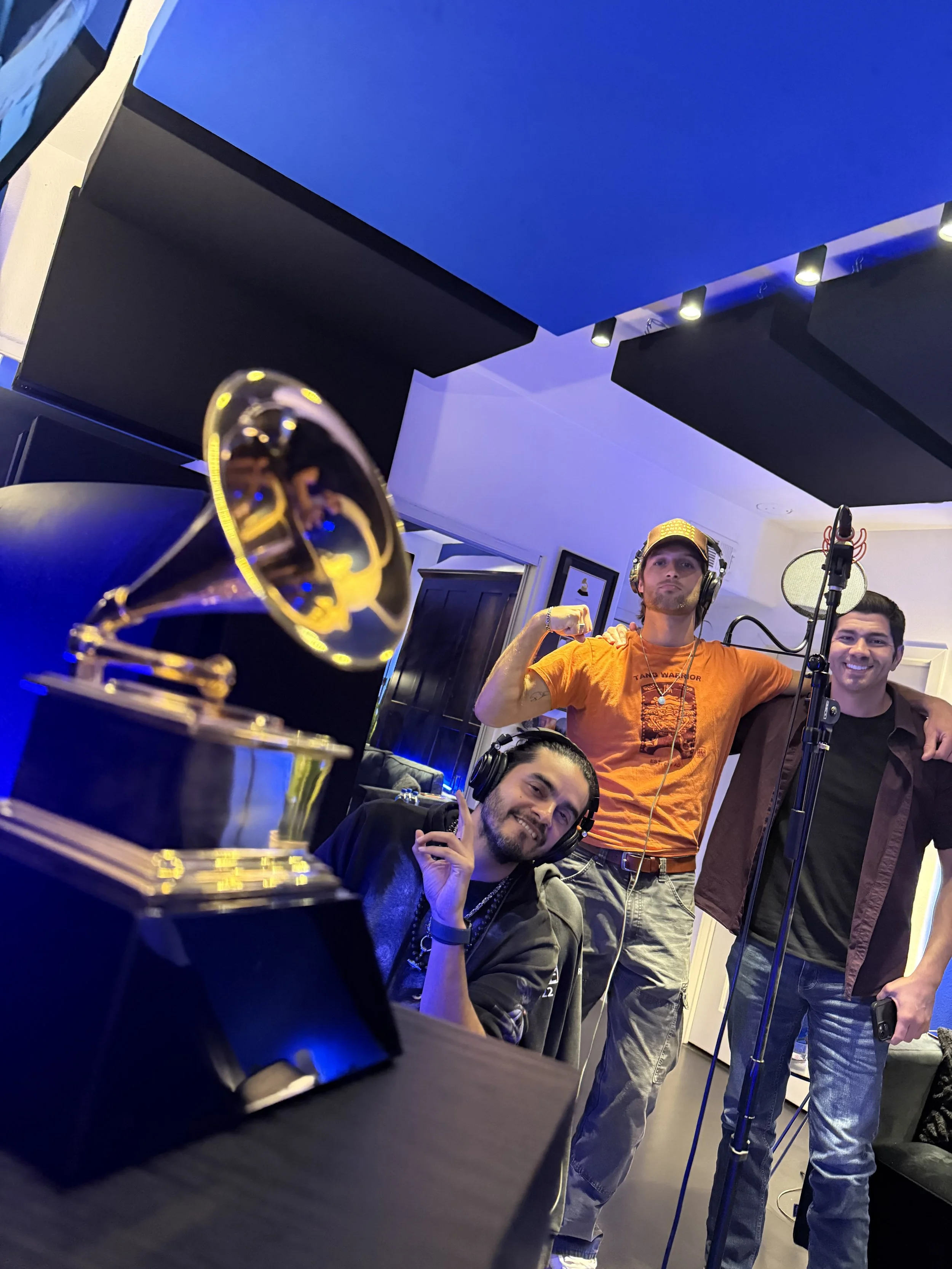 A group of people in a recording studio. One person is sitting at a table with a soundboard, smiling and making a peace sign. Two others are standing behind him, one flexing his arm and the other smiling. A microphone is in front of the group, and a 