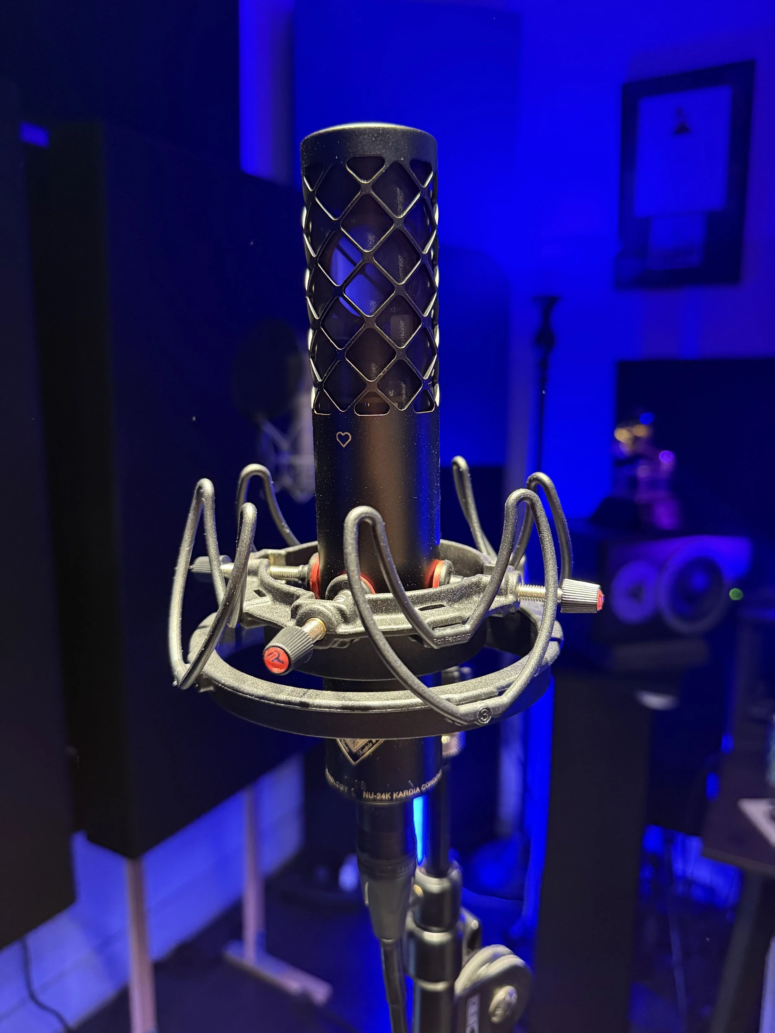 A studio microphone with a black mesh grille and a metal body mounted on a stand, illuminated with blue and purple lighting. The mic is the Nordic Audio Labs NU-24K mic.